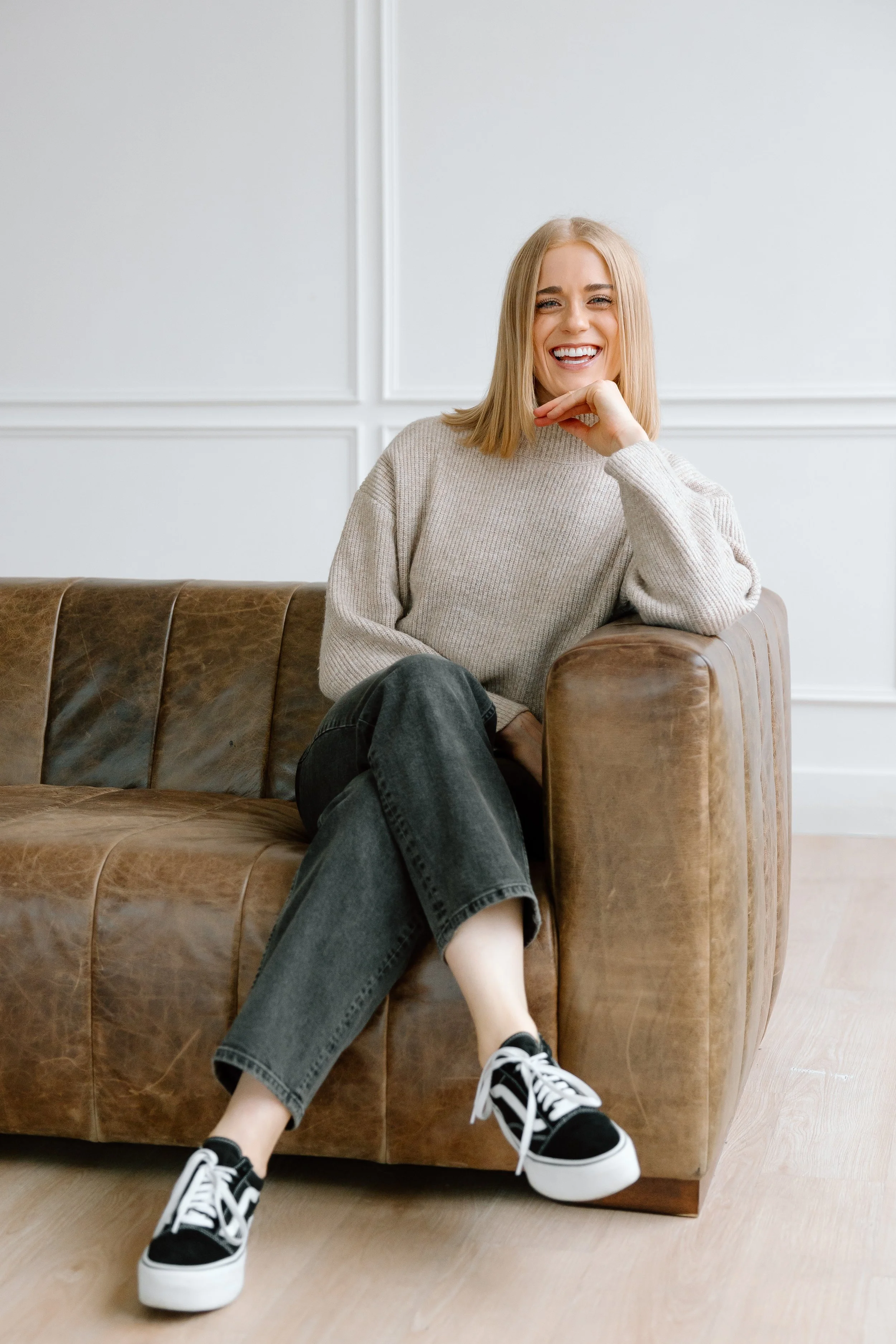 Smiling person sitting on a brown leather couch, wearing a gray sweater, black jeans, and black and white sneakers in a minimalistic room with white walls.