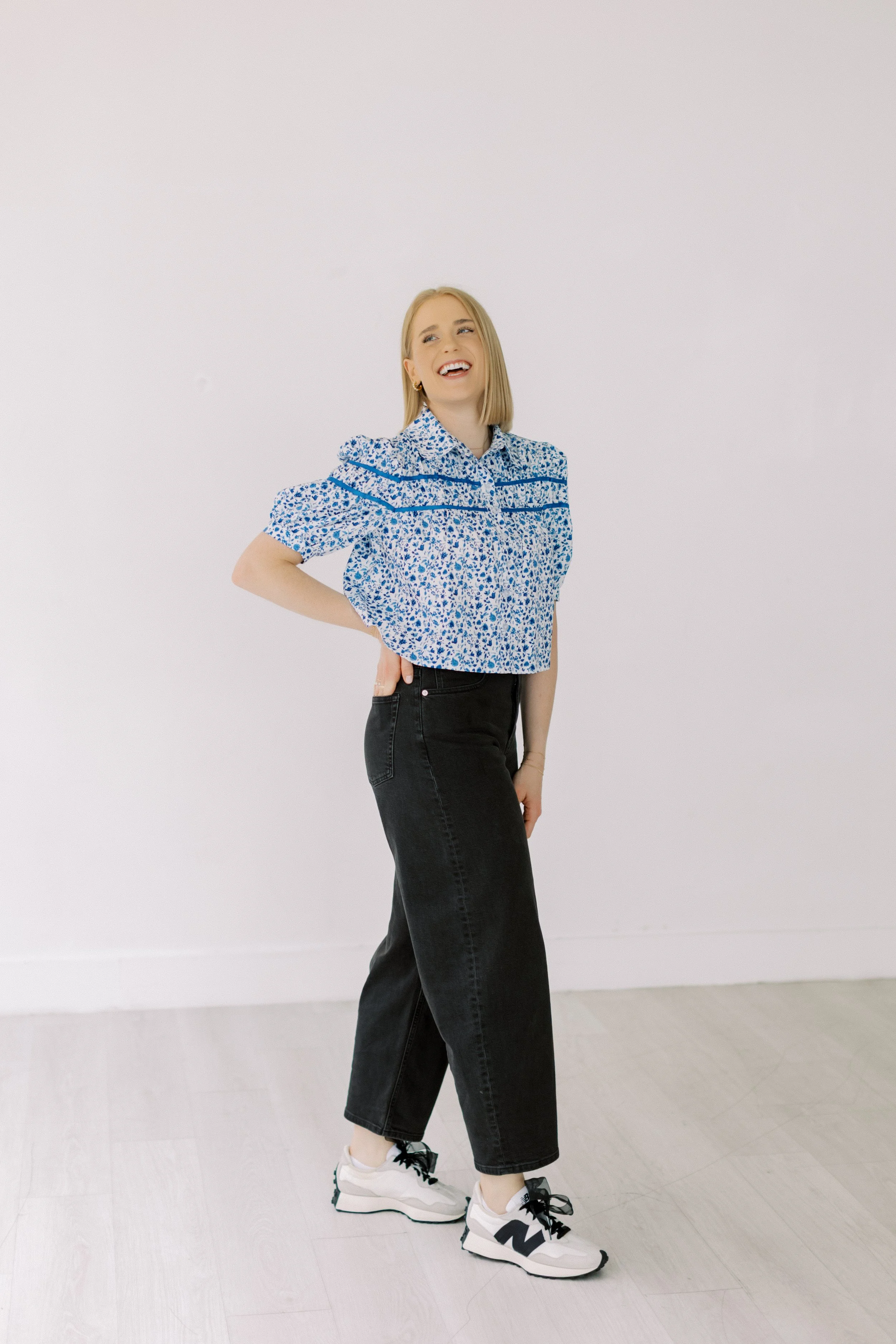 A young woman with blonde hair is standing against a plain white wall. She is wearing a blue and white patterned short-sleeve blouse, black wide-leg jeans, and white sneakers with black accents. She is smiling, looking upwards, with one hand on her hip and the other hanging by her side.
