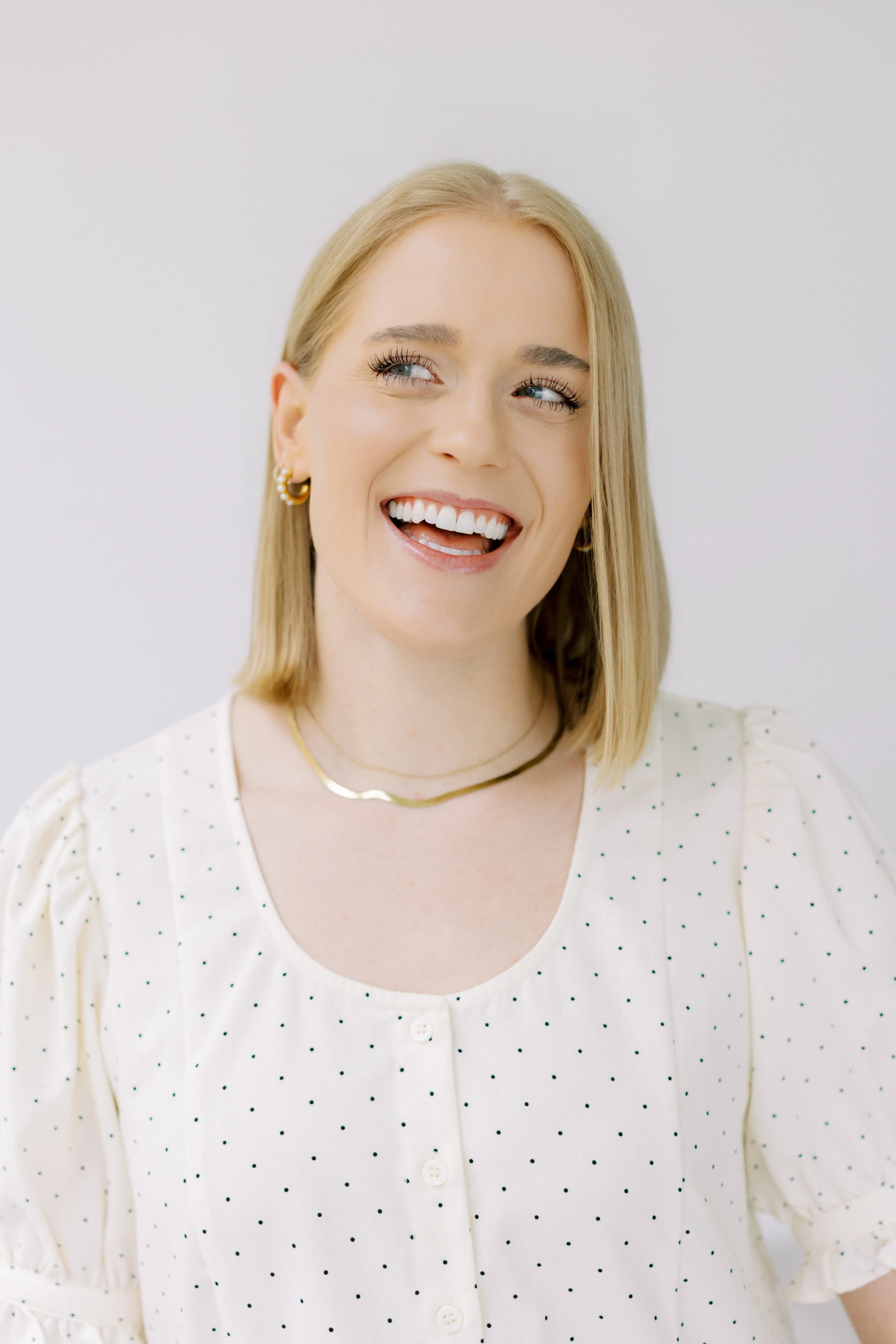 A woman with blonde hair and blue eyes smiling and looking slightly to her right, wearing gold earrings, a gold necklace, and a white blouse with small polka dots. Emily Rucker M.A. LPC. Houston Texas. Alive and Whole Wellness. Christian LPC.