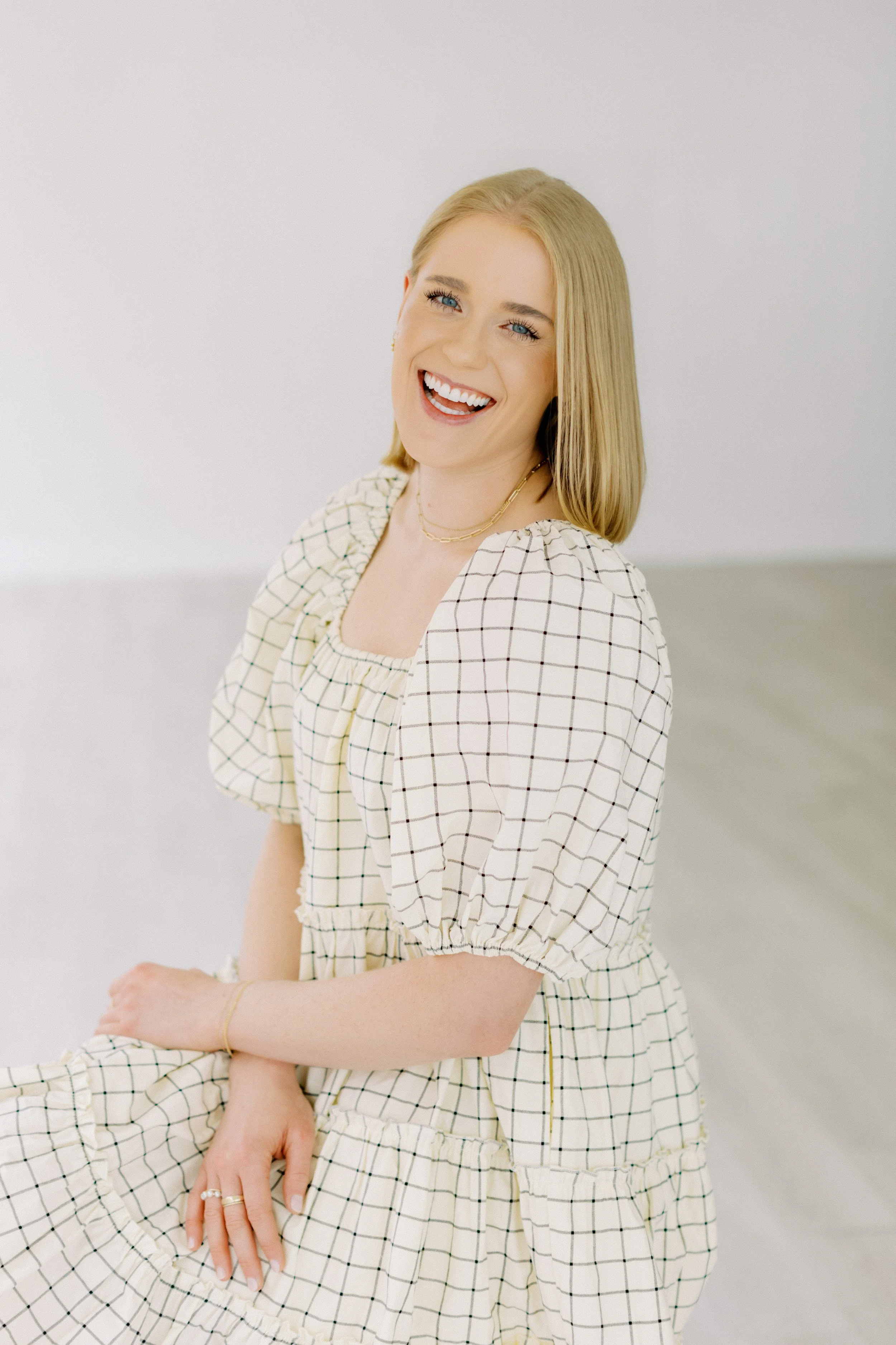 A smiling young woman with blonde hair and blue eyes, wearing a cream and black checked dress with puffed sleeves, sitting with her hands relaxed, in a minimal white background setting. Emily Rucker M.A. LPC. Houston Texas. Alive and Whole Wellness.