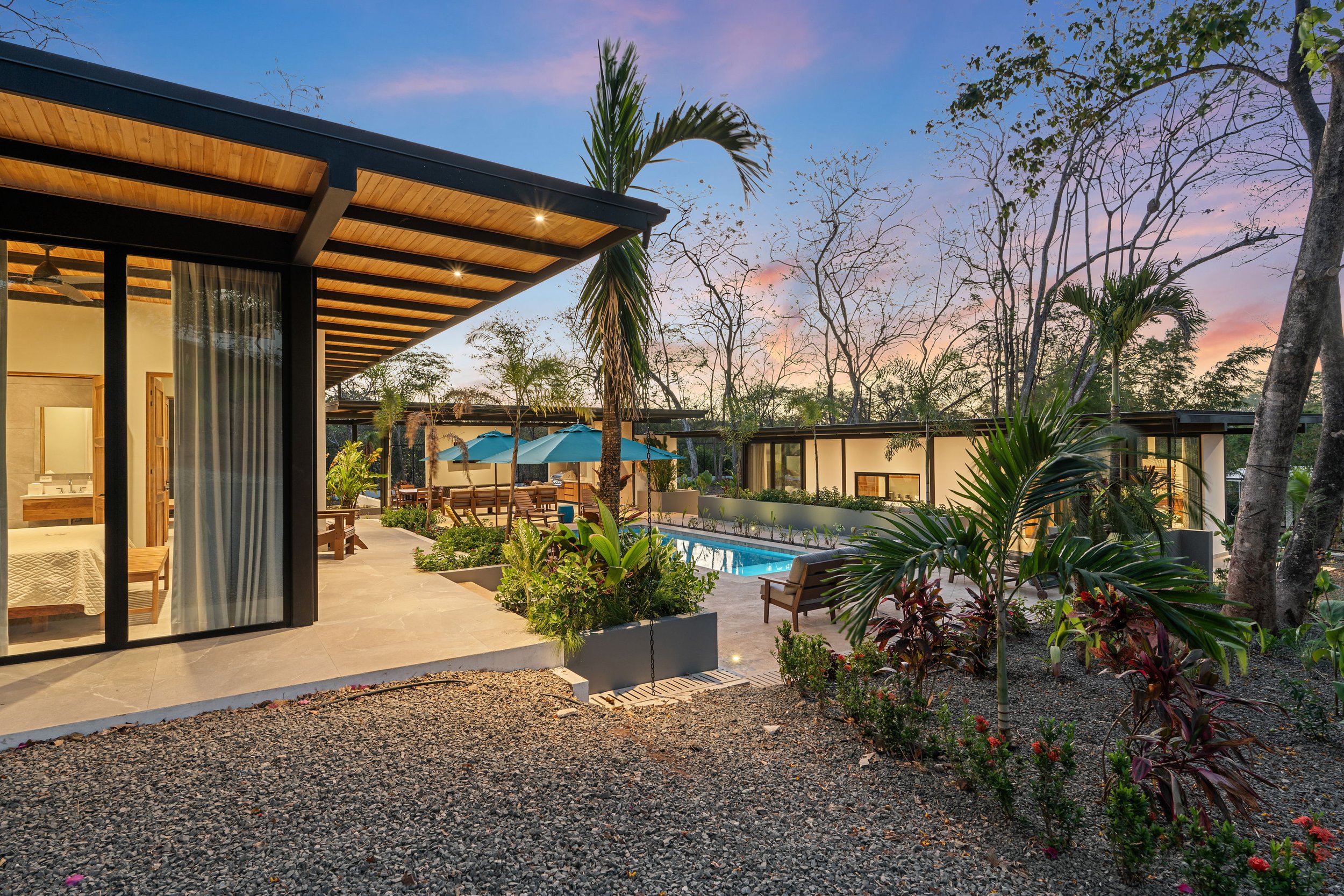 Modern outdoor patio with pool and plants, surrounded by contemporary buildings and tropical landscaping at sunset.