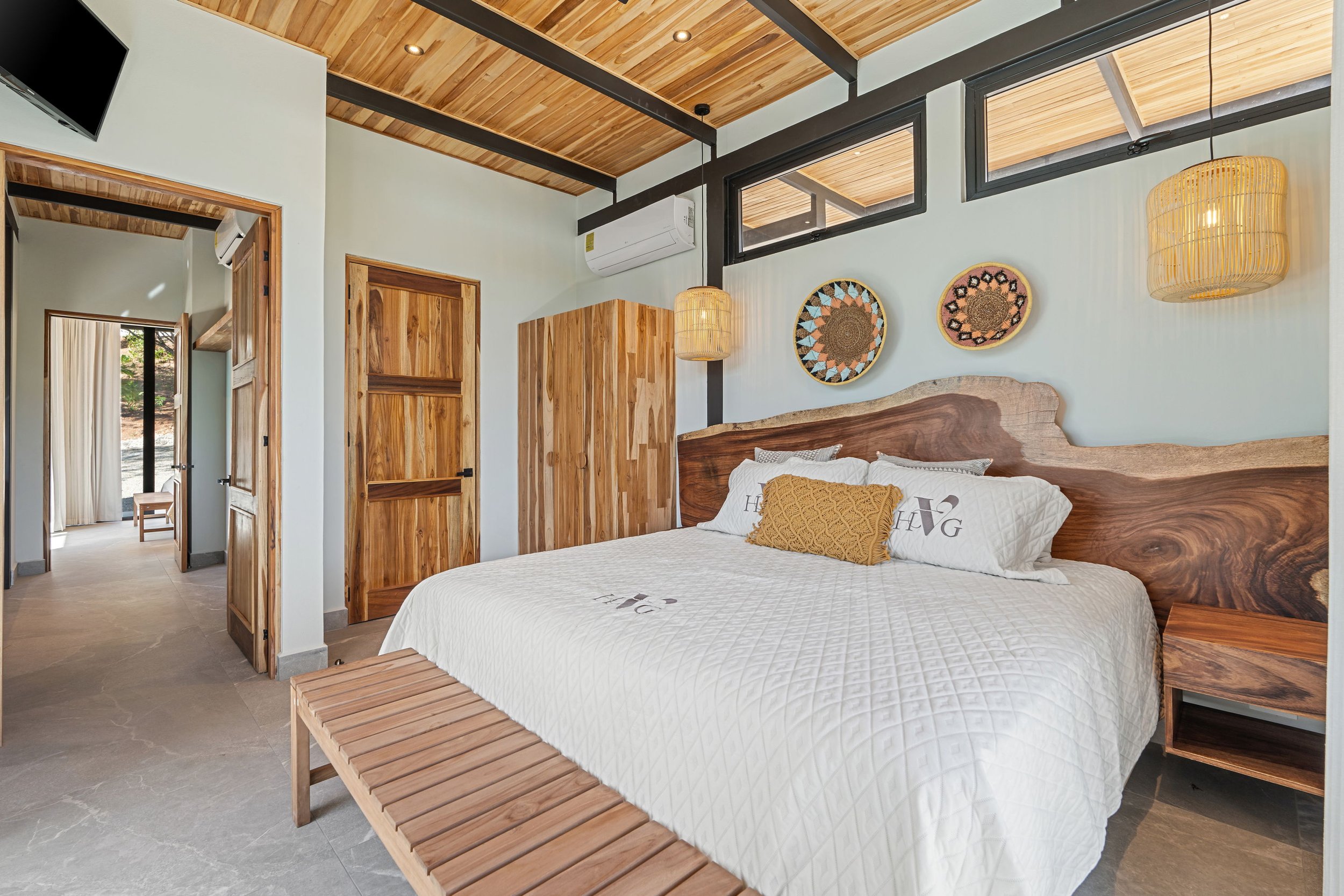 Modern bedroom with wooden ceiling and furniture, featuring a bed with a decorative headboard, wall hangings, and a hallway leading to a sunlit room.