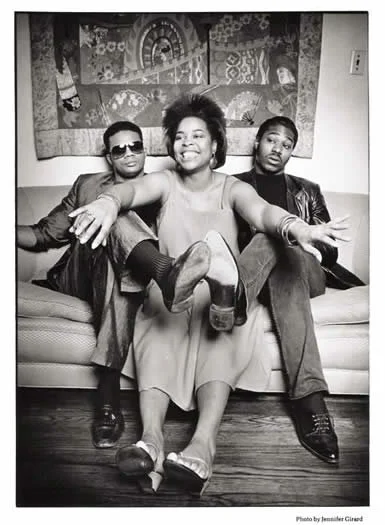 Three people sitting on a sofa, with the woman in the middle extending her arms and legs outward, smiling, in black and white photo.