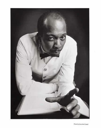 Black and white photo of a man in a tuxedo, leaning over a book, pointing towards the camera, with intense gaze.