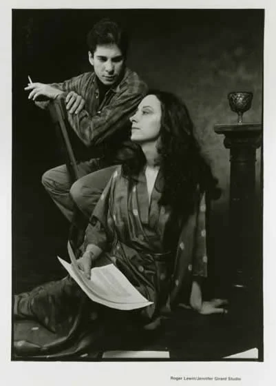 A black and white photograph of a man and a woman, with the woman sitting on the floor reading a book and the man standing behind her holding a cigarette. There is a pedestal with a decorative object on it nearby.