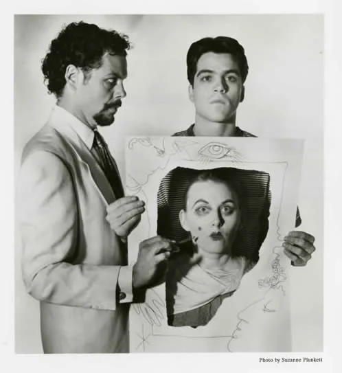 Two men, one with curly hair and beard, in a suit, and a young man with short hair, holding a framed portrait of a woman's face, with additional sketches and lines around the frame.