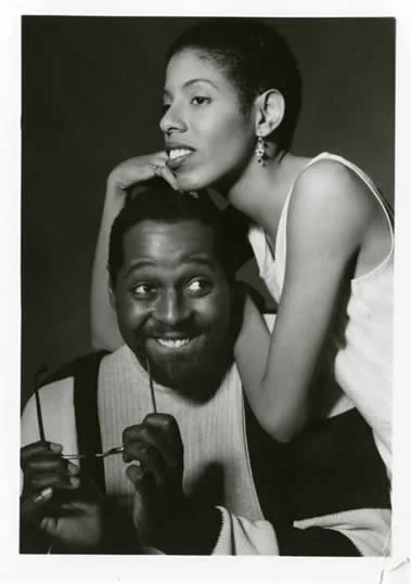 Black and white photo of a smiling man holding glasses in his hand and a woman leaning on his shoulder, both looking away from the camera.