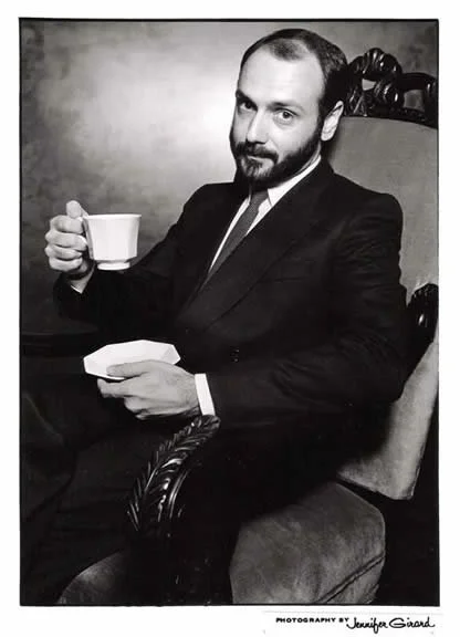 A man in a suit sitting in an ornate chair, holding a teacup and a small plate, with a serious expression in a black and white photograph.