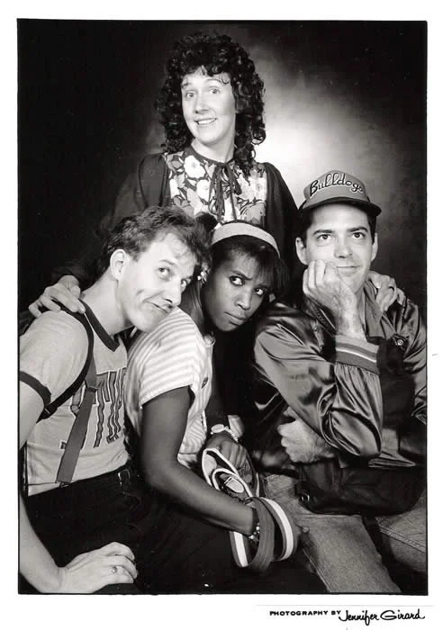 Black and white photo of four young people, three sitting in front and one standing behind them. The person at the back has curly hair and is smiling. The person on the left has short hair and is making a playful face. The person in the middle has da