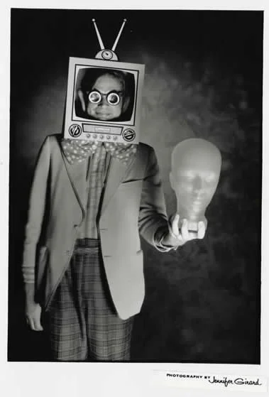 Person dressed in a suit with a vintage television set as a head, wearing glasses, holding a mannequin head, in black and white.
