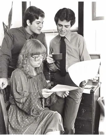 Three young people, two men and one woman, gathered around looking at documents in an office setting, with a potted plant and window in the background.