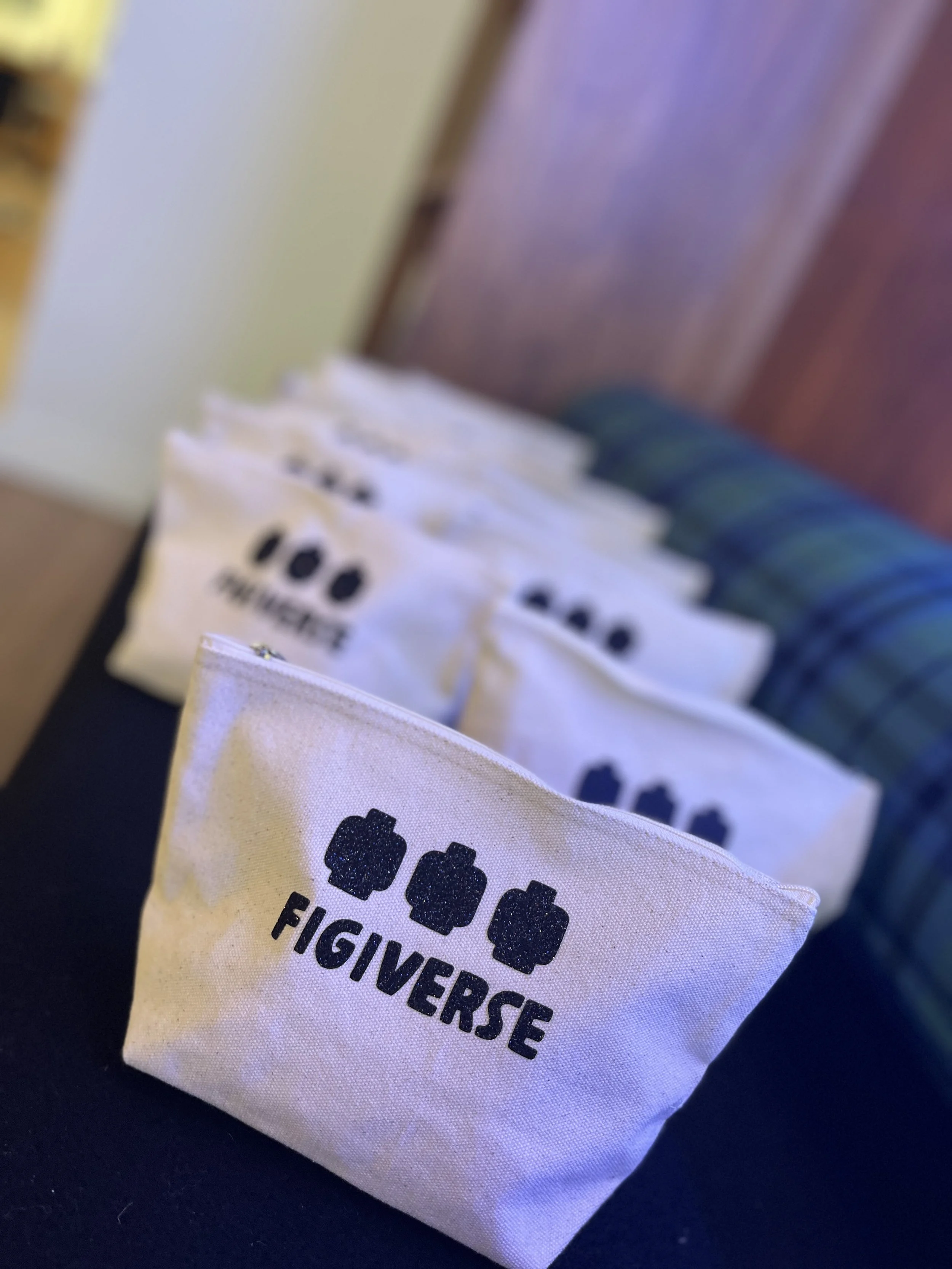 A row of white pouches with black logo and text on a black surface. The pouch in foreground displays three stylized figures above the word 'FIGIVERSE'.