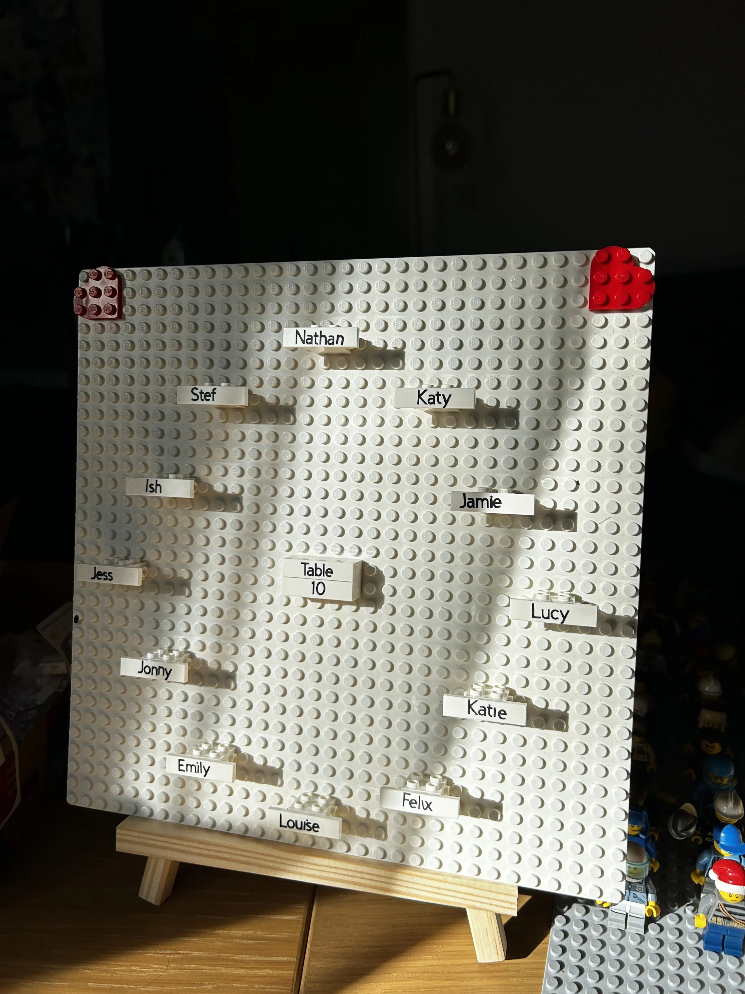 LEGO-style board with small white labels with names attached to a grid of white circular studs, some labels are 'Nathan', 'Katy', 'Jamie', 'Lucy', and 'Katie'. There is a small rectangular block labeled 'Table 10' in the center.