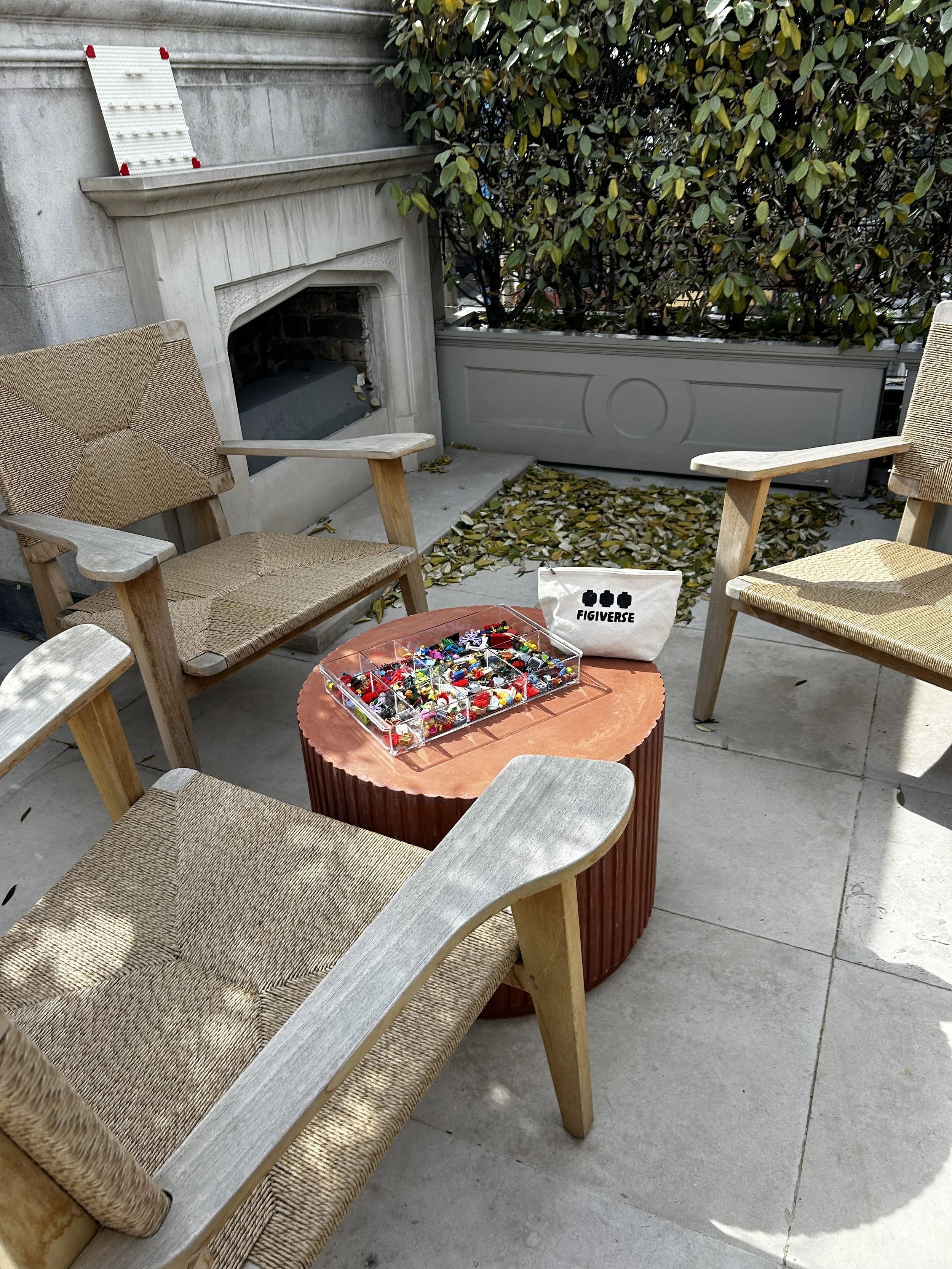 Outdoor patio with four woven armchairs surrounding a round wooden table with divided storage box filled with colorful sewing pins. A white bag labeled "FIGIVERSE" is on the ground near a black stone fireplace and leafy bushes in the background. Fall