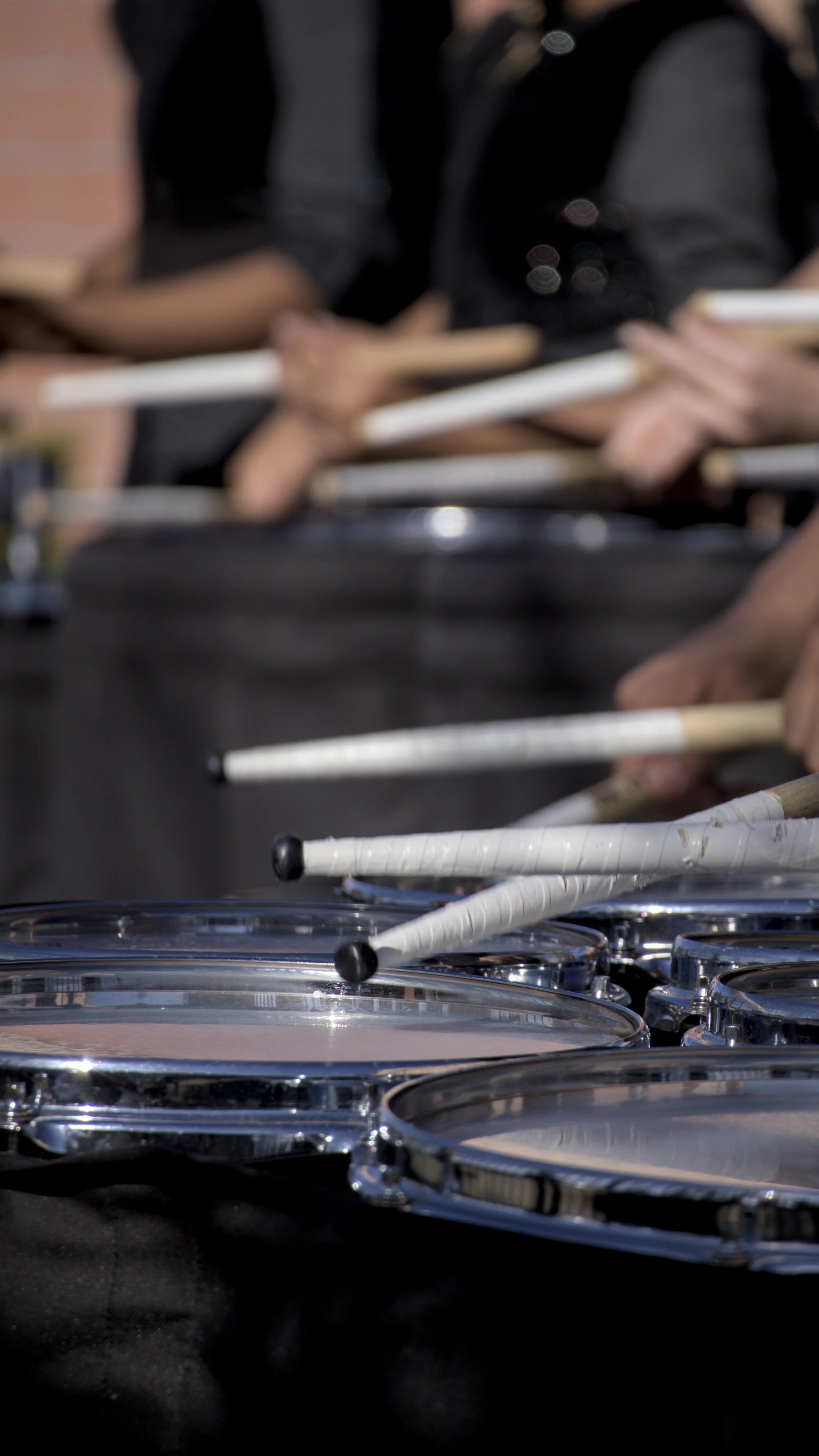 Close-up of drummers playing snares with drumsticks, wearing dark clothing.