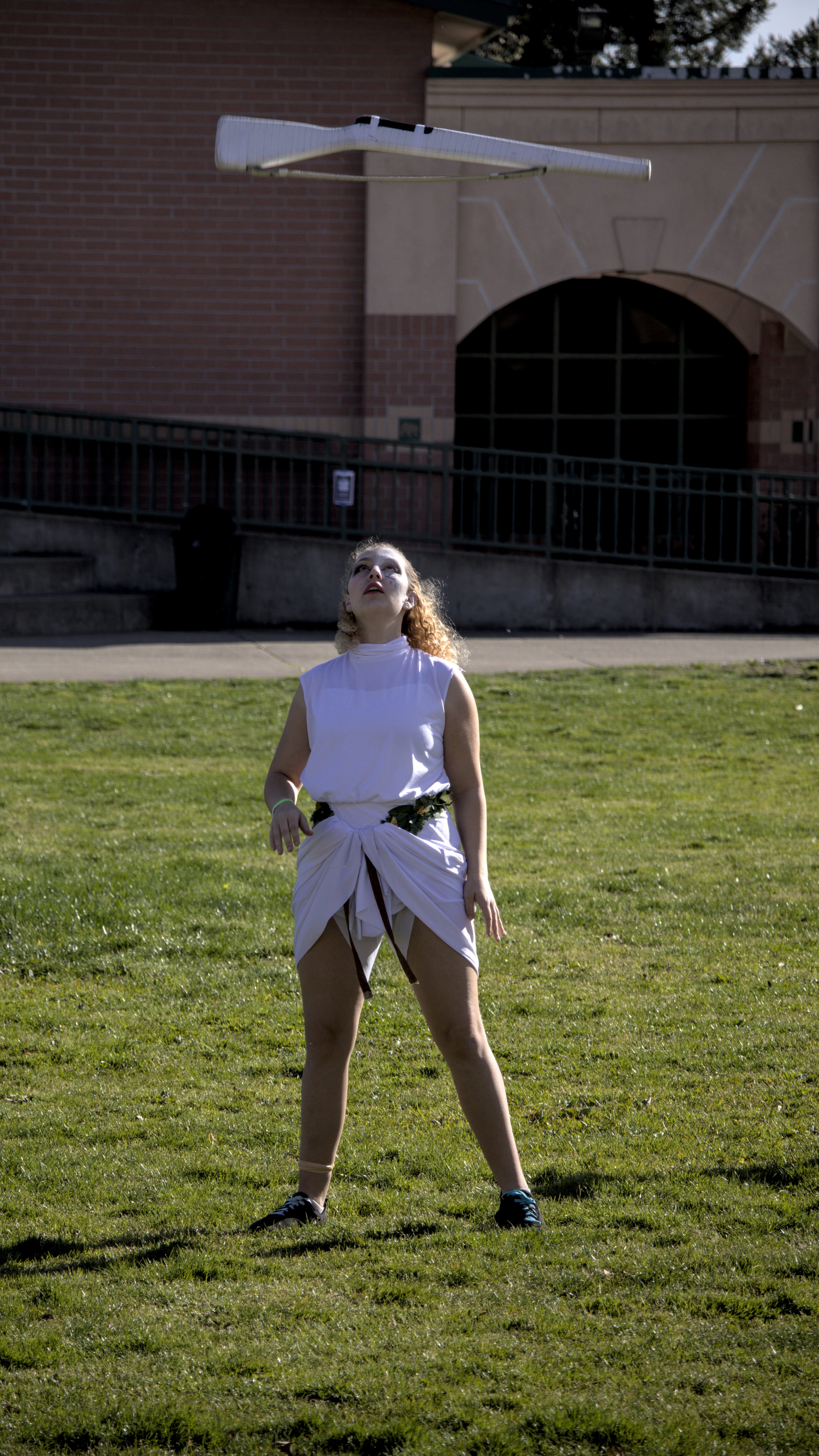 Person outdoors on grass, wearing a white outfit with a leaf belt, looking up at an object hovering above them.