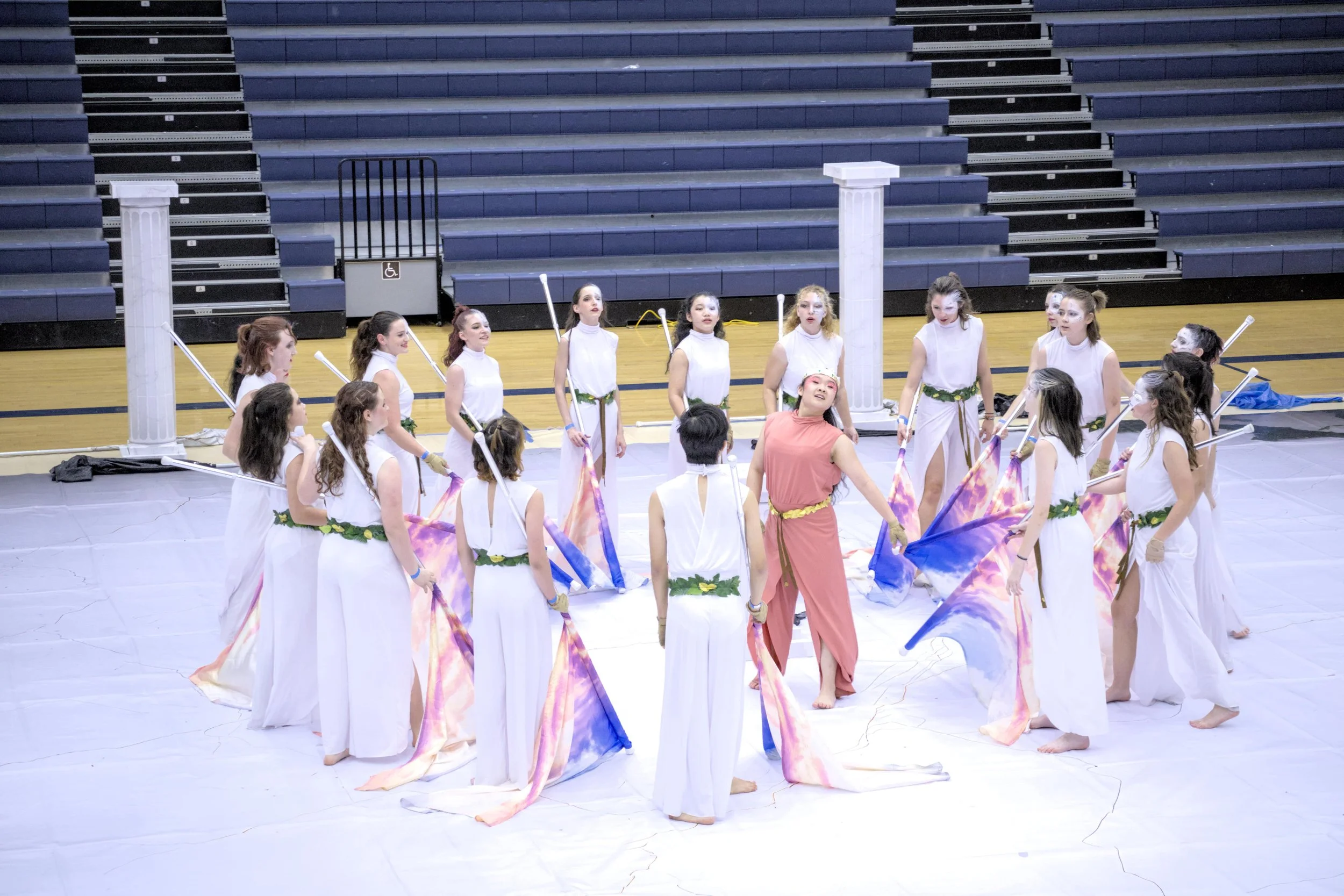 Group of performers in a gymnasium dressed in white and pink costumes, holding colorful flags, with two white columns in the background.