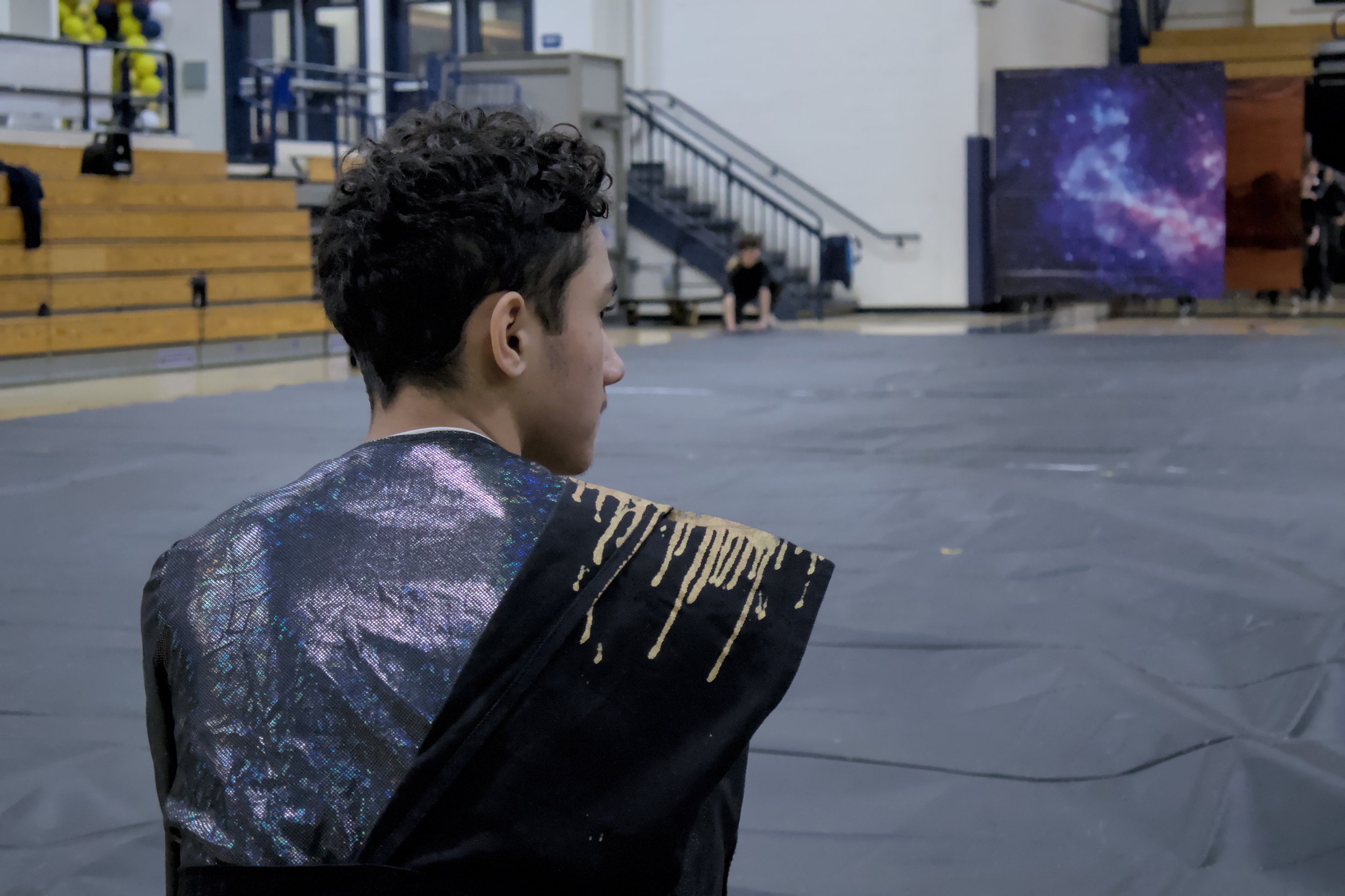 A person in a gym setting wearing a shiny, reflective jacket with gold accents, facing away from the camera. The background includes bleachers and a cosmic-themed backdrop.