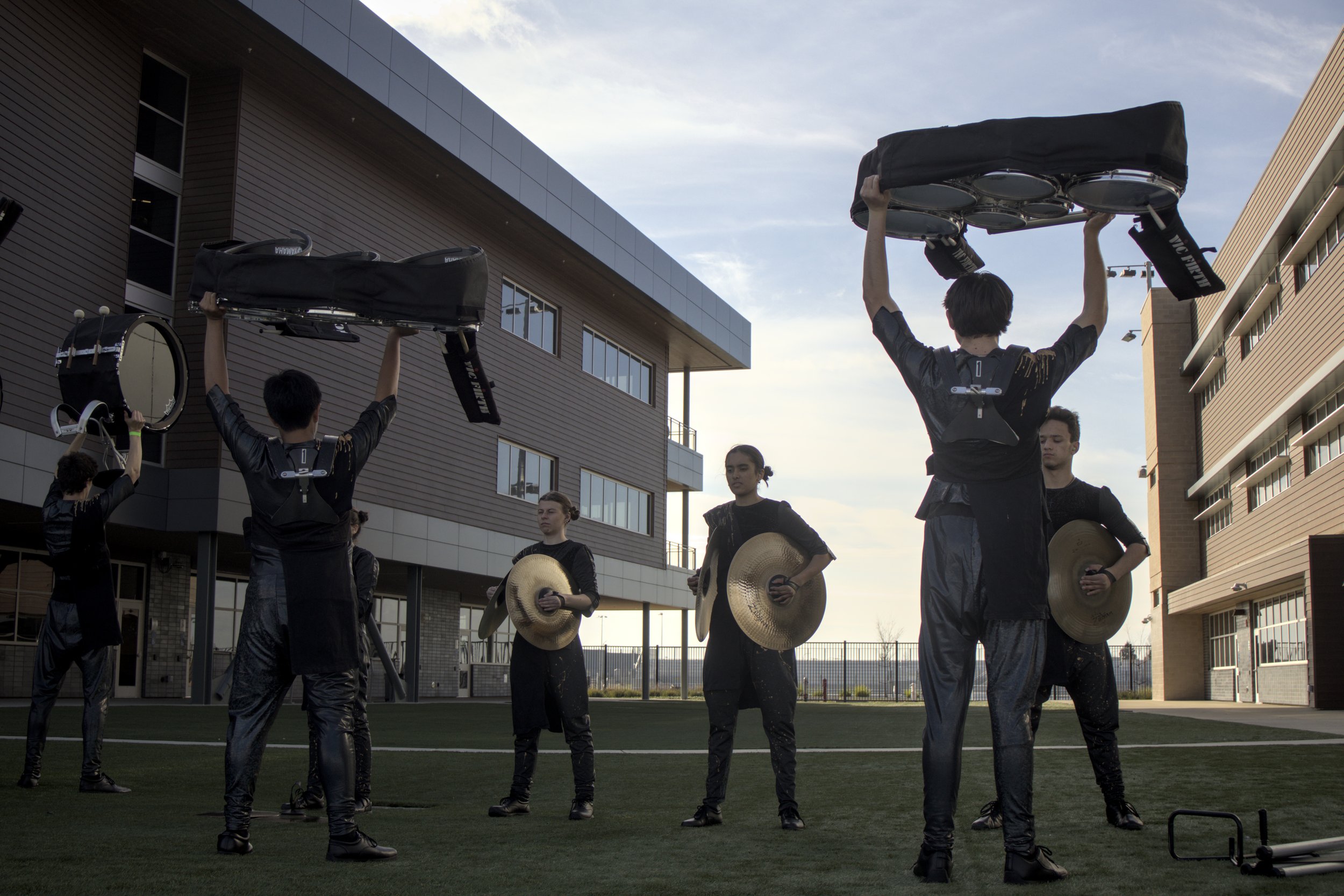 Marching band percussion section with musicians holding drums and cymbals outdoors near buildings.