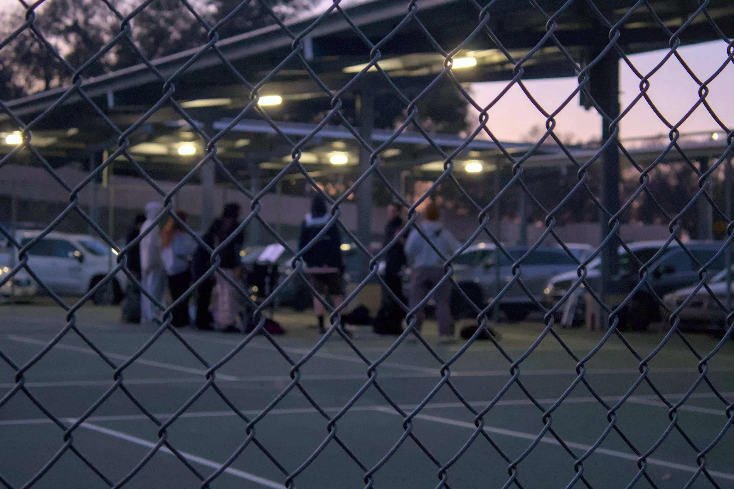 View through chain-link fence of people gathered on a sports court with cars and a covered parking area in the background.