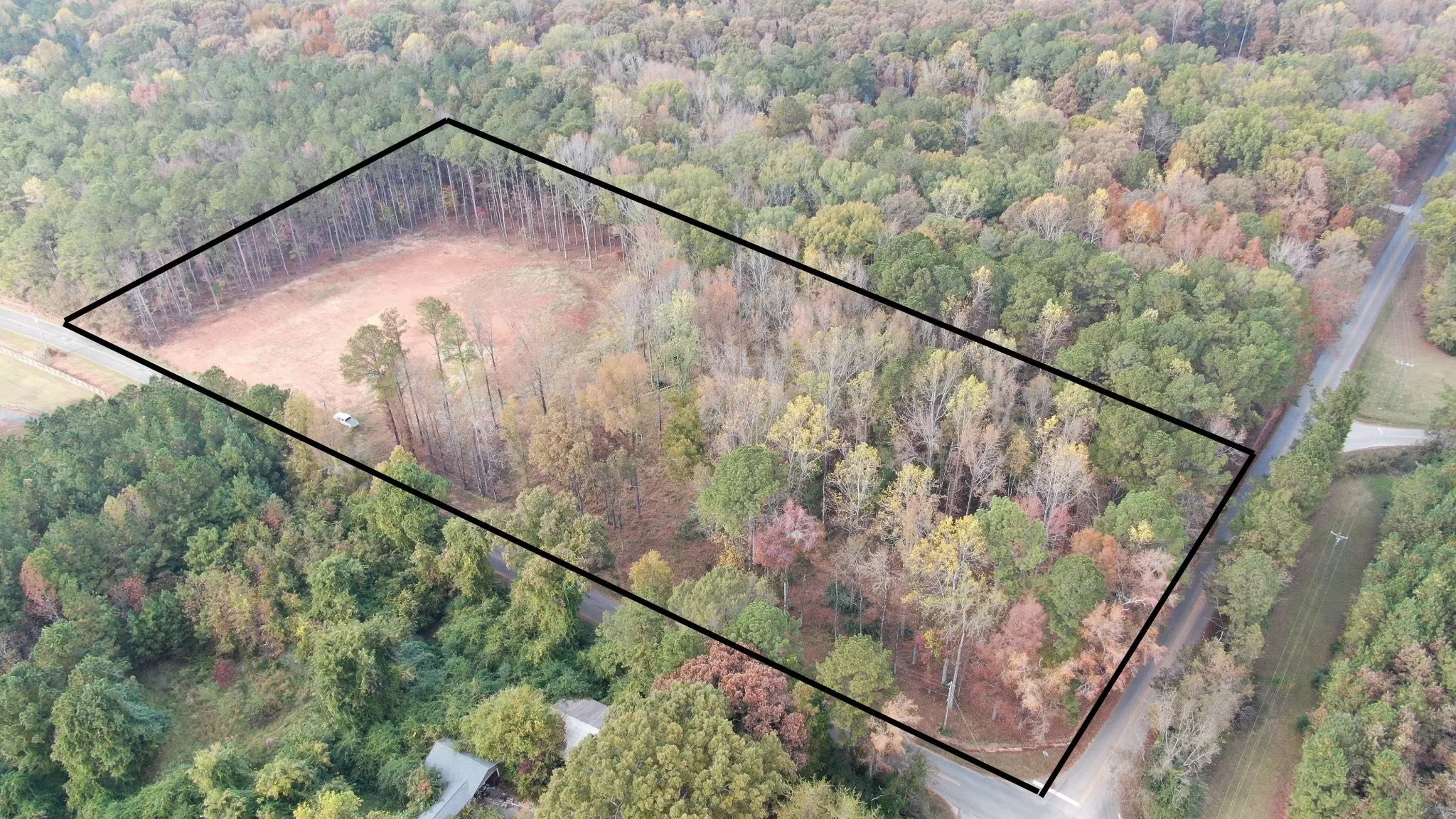 Aerial view of a rectangular plot of land outlined in black, surrounded by a dense forest with a road on one side. There is a small cleared area with no trees and a car parked nearby.