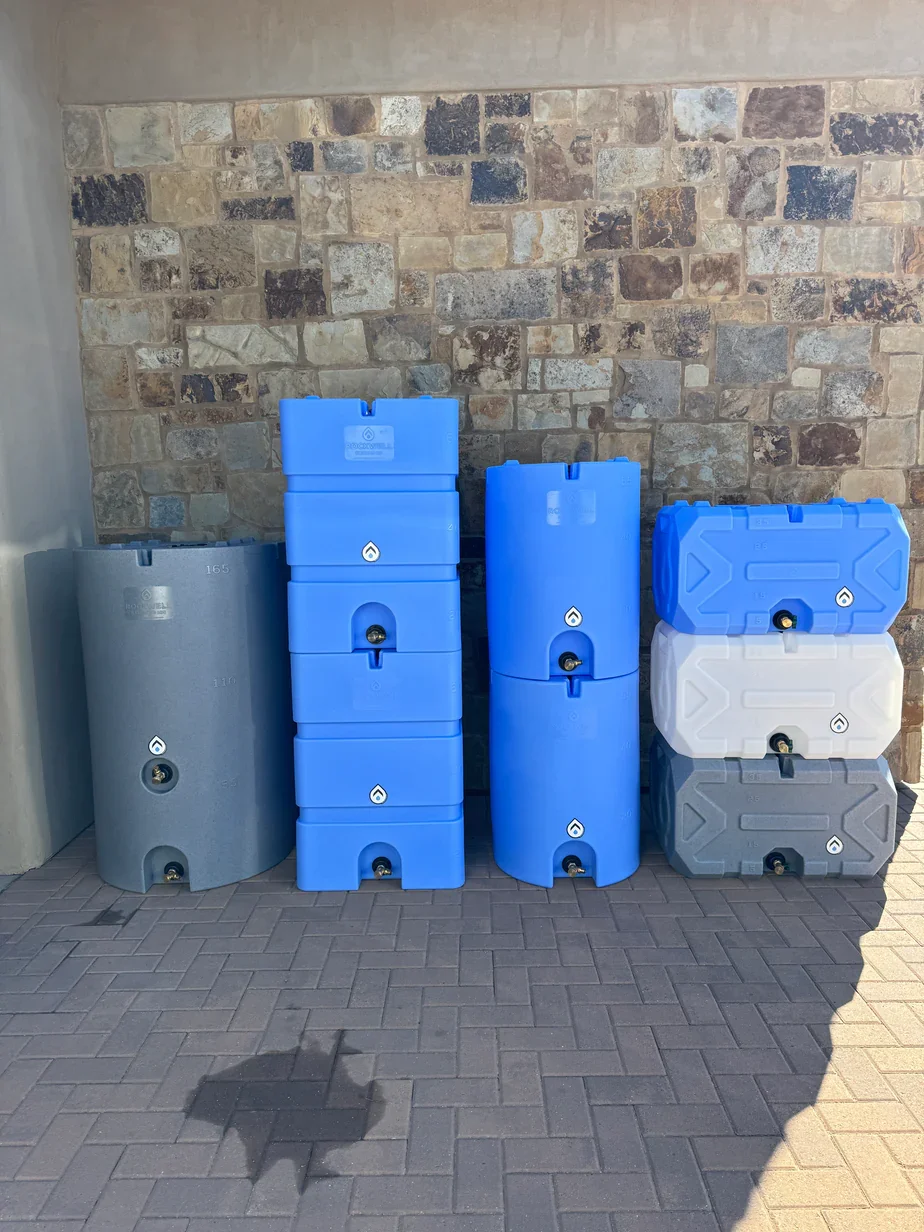 Stacked water storage tanks in gray, blue, white, and gray colors against a brick wall on pavement.