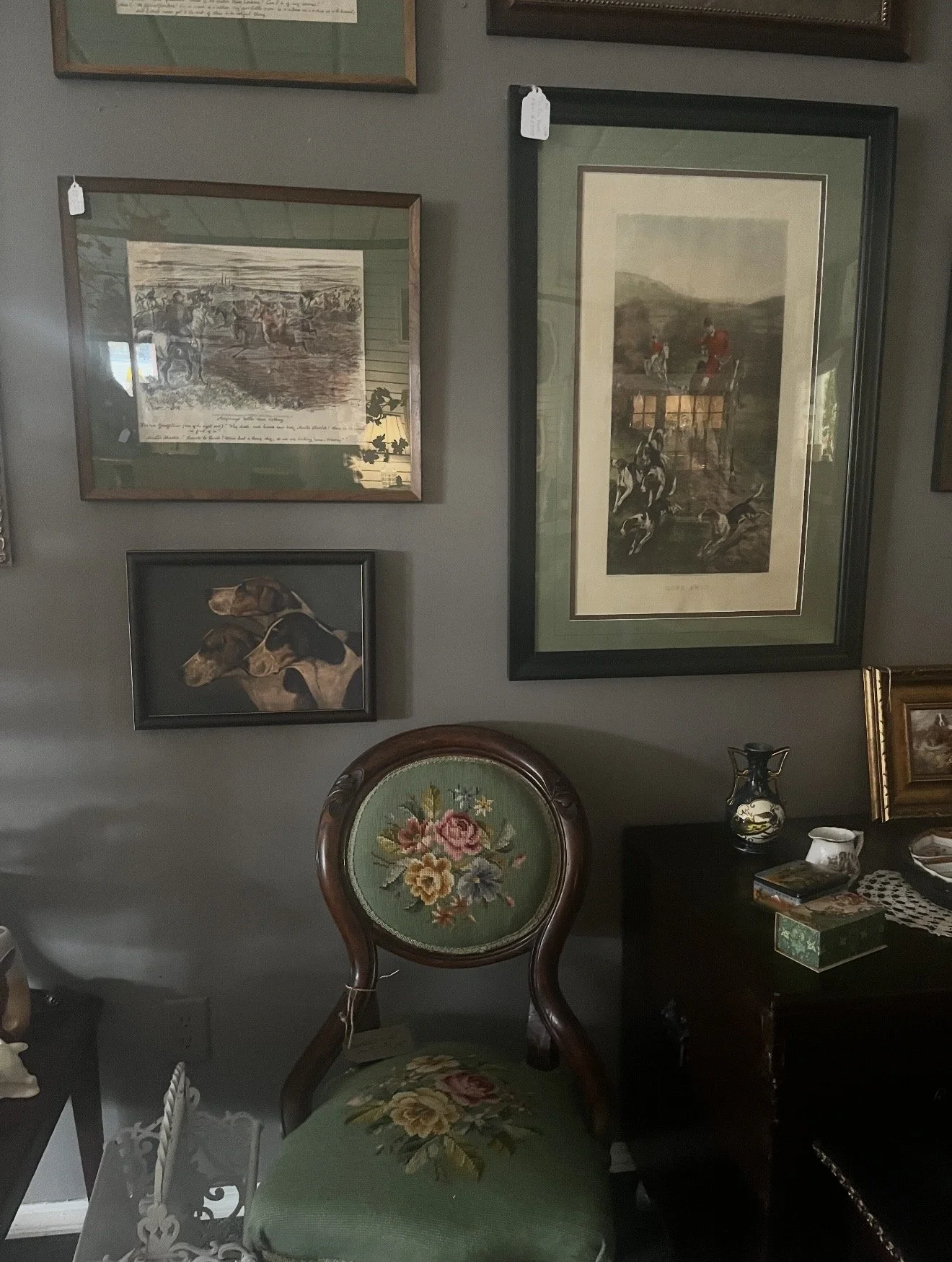 A living room with gray walls decorated with multiple framed pictures and paintings. There is a vintage green upholstered chair with a floral design on the backrest and seat, placed in front of a black side table that holds decorative items, includin