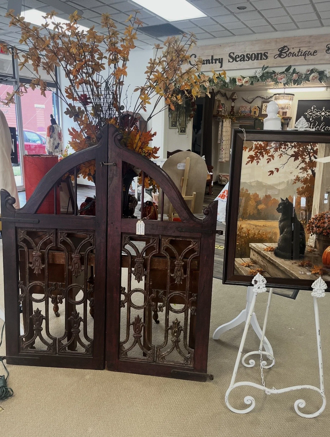 A display of fall-themed home decor inside a store, featuring a wooden decorative gate, artificial autumn leaves, a framed painting of a black cat sitting outdoors, and various decorative objects.