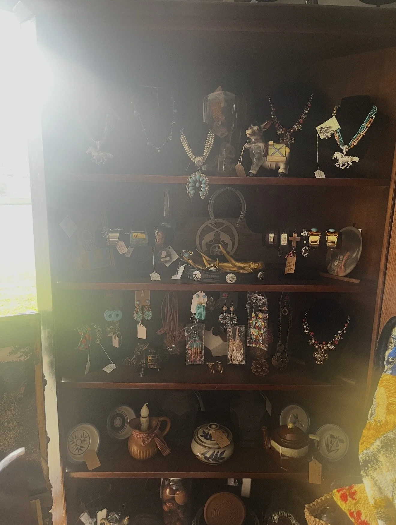 A display shelf filled with various jewelry and decorative items such as necklaces, rings, earrings, and small pottery vessels. The shelves also contain small figurines, a miniature toy car, and other miscellaneous collectibles.