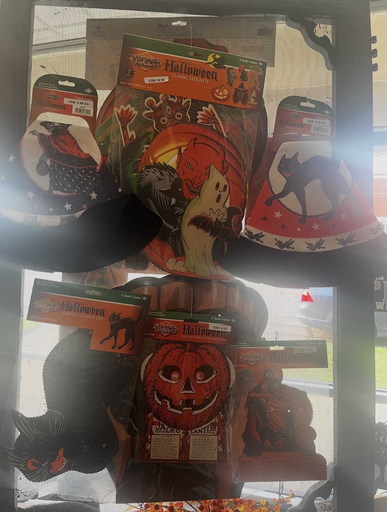 Various Halloween-themed Halloween hats with decorations, including pumpkin faces, black cats, witches, and bats, displayed on hooks behind a window screen.