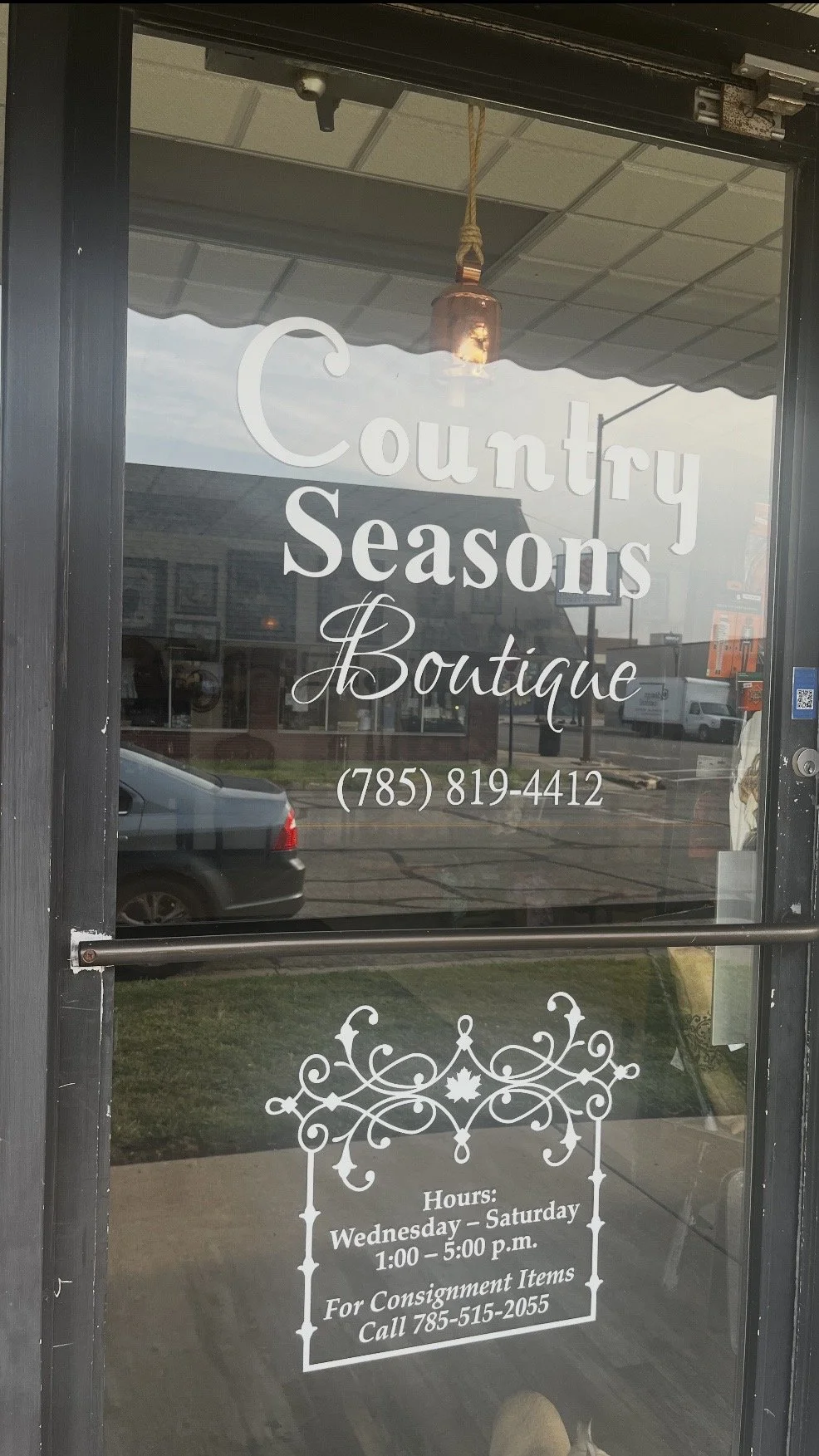 Glass door with white text displaying the name 'Country Seasons Boutique,' phone number, hours from Wednesday to Saturday 1:00 to 5:00 p.m., and a note about consignments along with a decorative design.