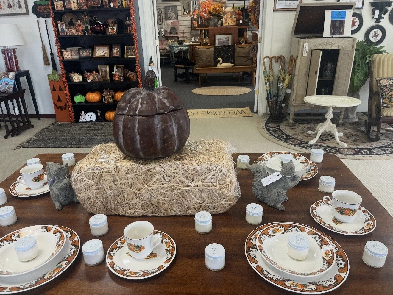 A wooden dining table with Halloween-themed dinnerware, small candles, and a large pumpkin centerpiece, set in a decorated room with shelves of pumpkins and Halloween decor in the background.