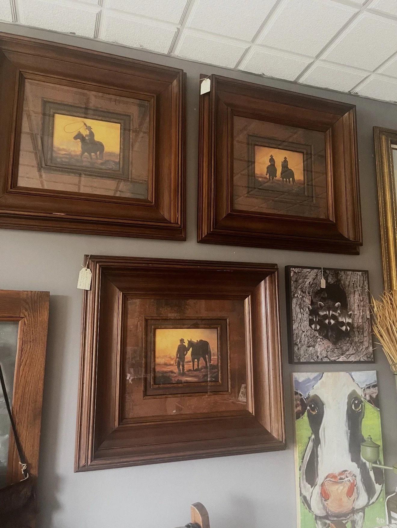 Wall of framed pictures, including three with Western cowboy and horse themes, and a color illustration of a cow and calf.