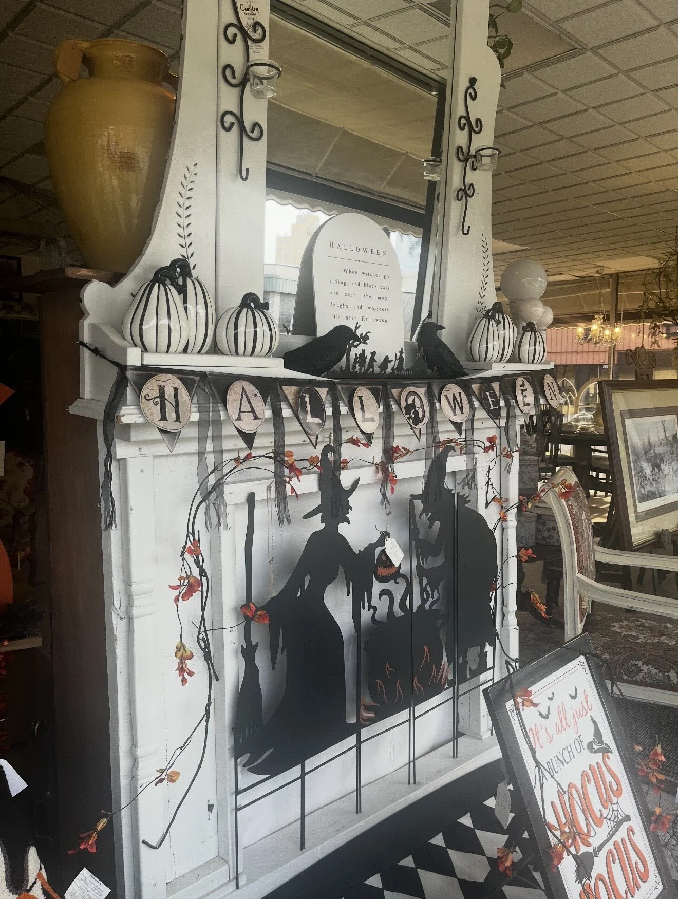 Store display with Halloween decorations, including a white fireplace mantel painted with black silhouettes of witches, a black cat, and a cauldron, topped with white pumpkin ornaments, a mirror, and a large yellow vase on the side, with a Halloween 