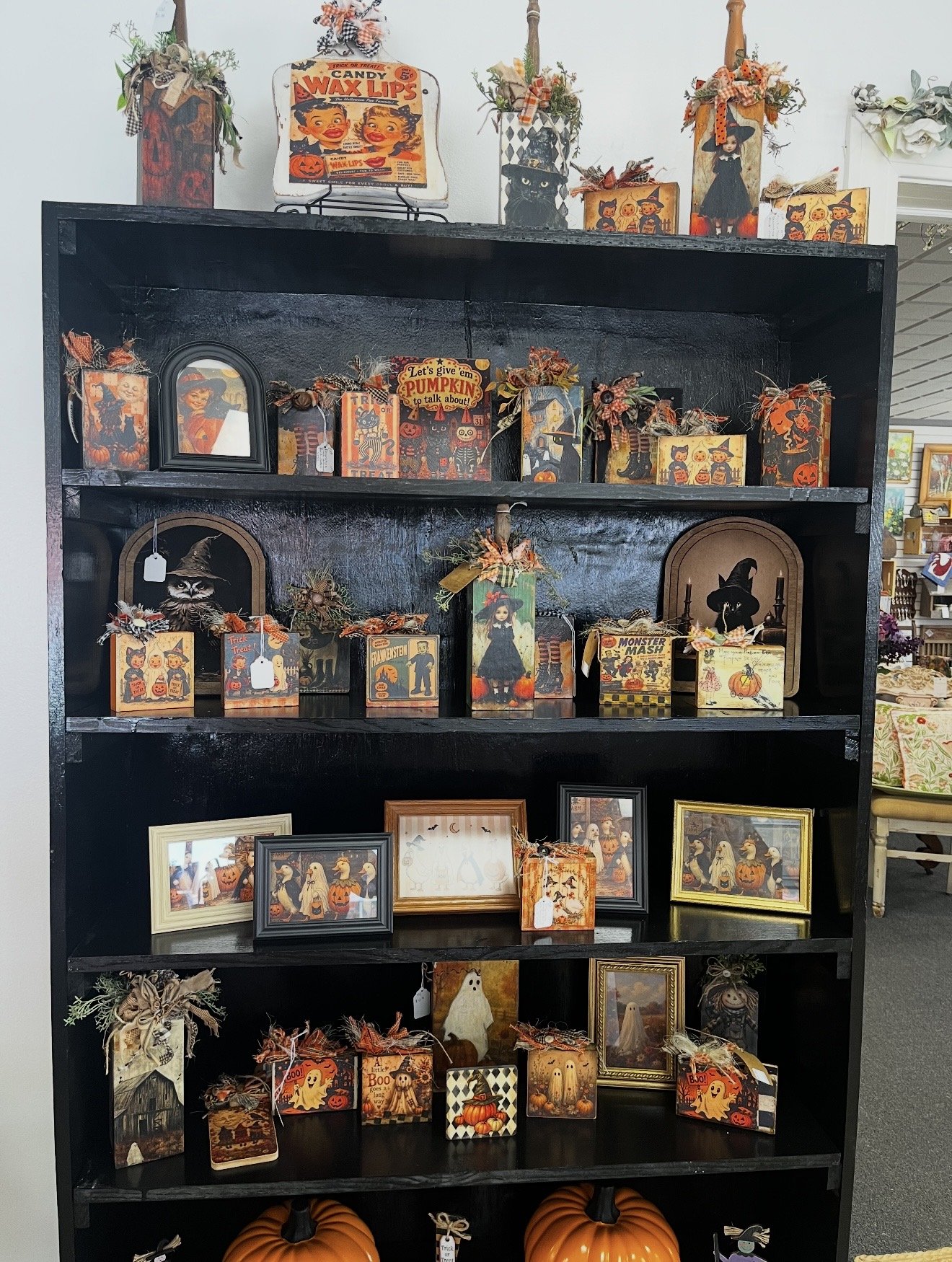 Black bookshelf decorated with various Halloween-themed decorations and picture frames. The top shelf displays vintage-style signs and small floral arrangements with ribbons. The middle shelves contain framed artwork, small signs, and decorative item