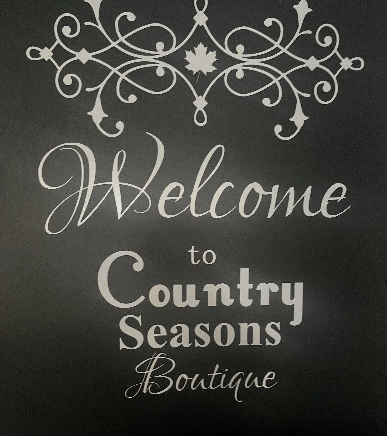 Decorative sign with the words 'Welcome to Country Seasons Boutique' written in elegant white script on a dark background, adorned with ornate white scrollwork and a maple leaf design at the top.