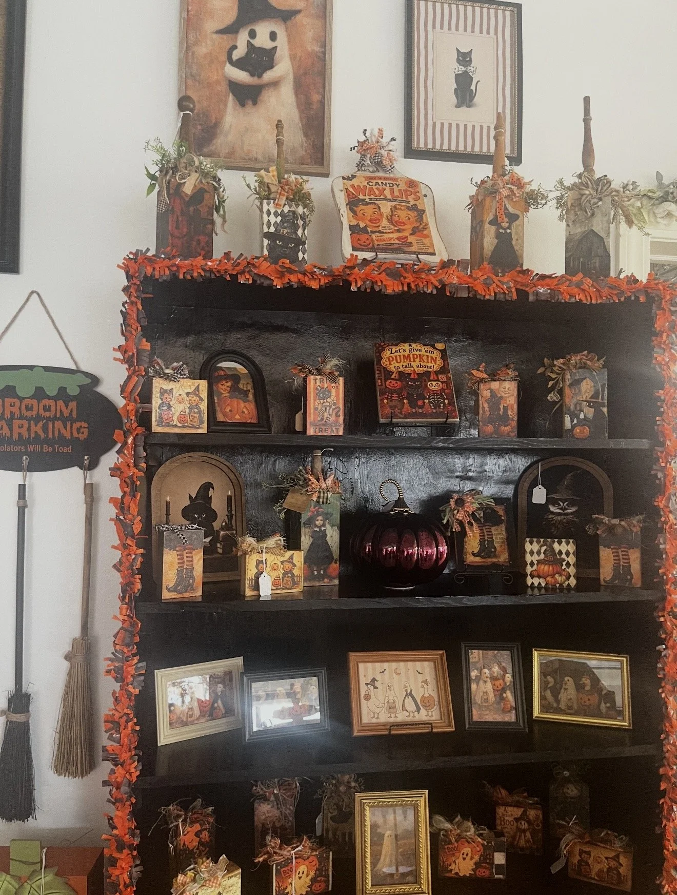 Display shelf decorated for Halloween with various themed decorations, framed pictures, and orange and black accents, including artwork of black cats and witches, and Halloween-themed signs and cards.