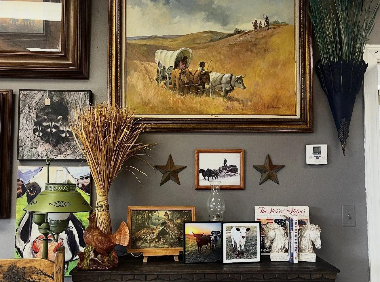 Wall decorated with various framed pictures and decor, including a large landscape painting of oxen pulling a wagon, smaller images of farm animals, a raccoon image, a bird sculpture, star-shaped decorations, a lamp, and a glass oil lamp.