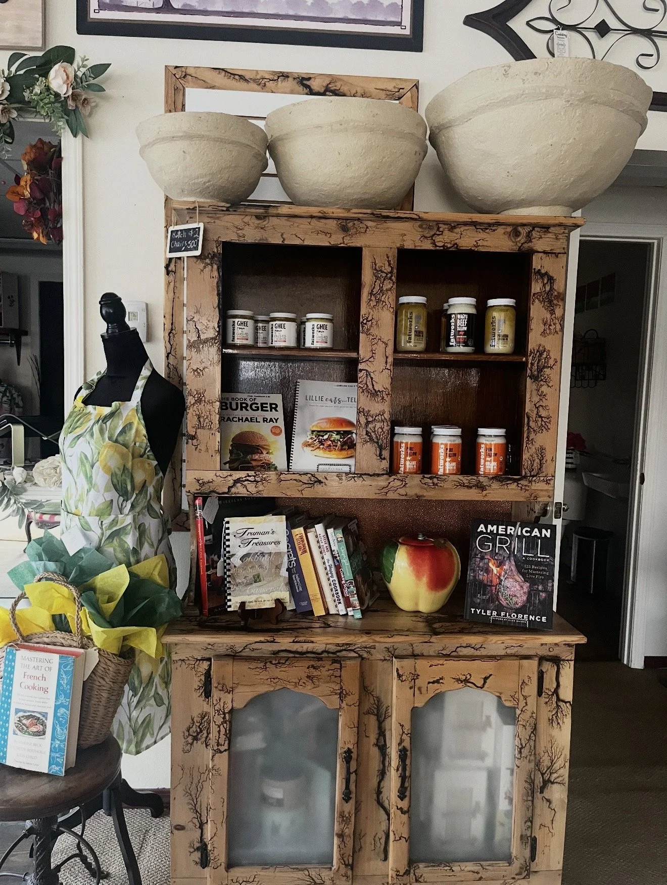 A wooden hutch decorated with a cracked paint finish, displaying various cookbooks, a large ceramic apple, and jars of spices or condiments. On top of the hutch, there are three large stone bowls. To the left, a mannequin dressed in an apron stands b