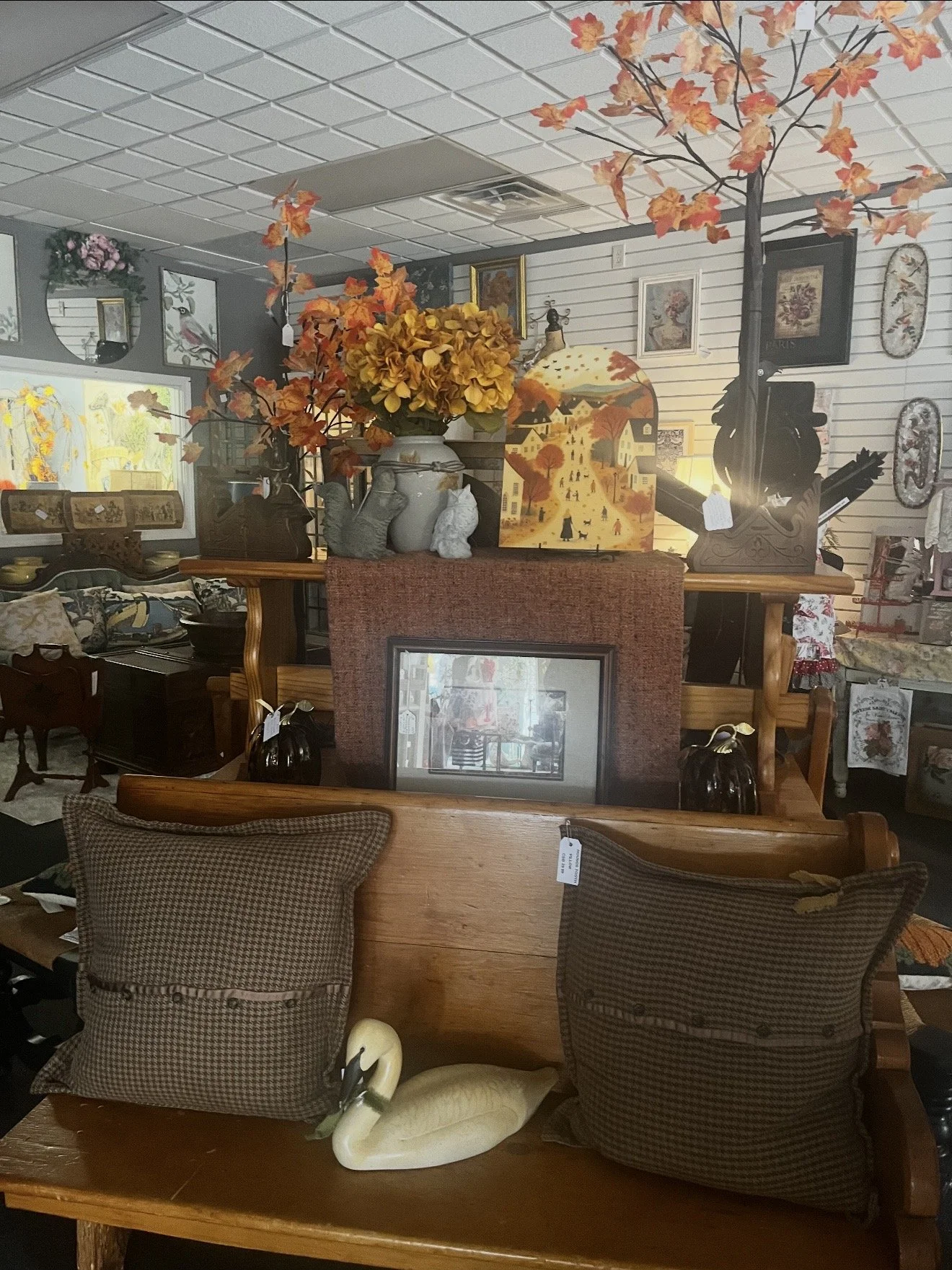 Interior of a vintage-style store with decorative items, floral artwork, and wooden furniture, including chairs with patterned cushions, a swan figurine, and framed pictures on the wall, with autumn-themed paper trees and market tags visible.