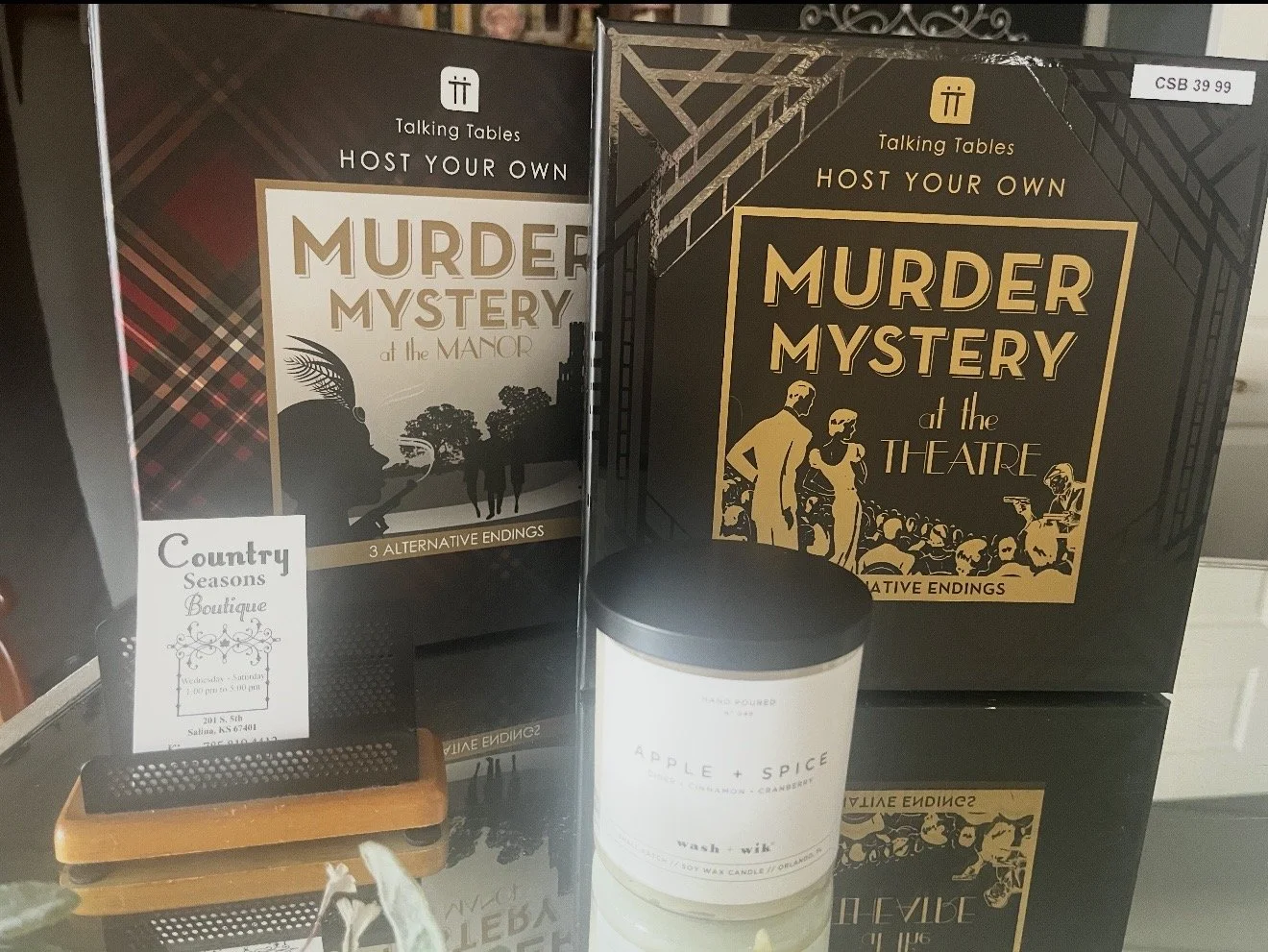 Two boxed murder mystery games titled 'Host Your Own Murder Mystery at the Manor' and 'Host Your Own Murder Mystery at the Theatre' with three alternative endings, along with a white candle labeled 'Apple + Spice' and a small sign for Country Seasons Boutique, displayed on a table.