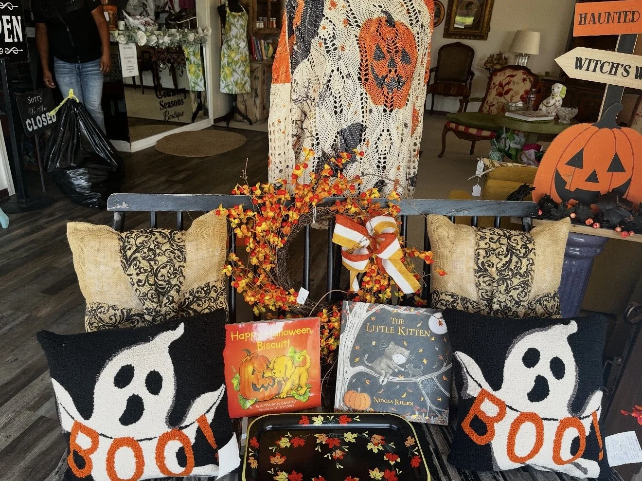 Autumn-themed Halloween display with pumpkin-shaped pillows, books, a decorative wreath with orange and yellow berries, and Halloween decorations including a large pumpkin with a carved face and a witch's hat sign in a cozy store setting.