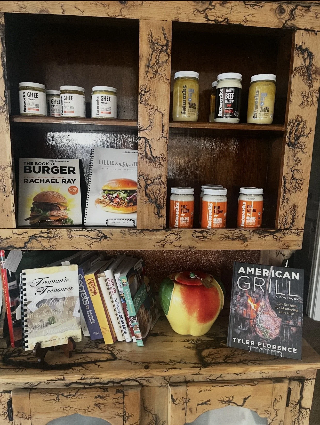 Wooden bookshelf containing jars of ghee and cooking oils, cookbooks including 'The Book of Burger' by Rachael Ray, local cookbooks, a decorative apple, and a book titled 'American Grill' by Tyler Florence.