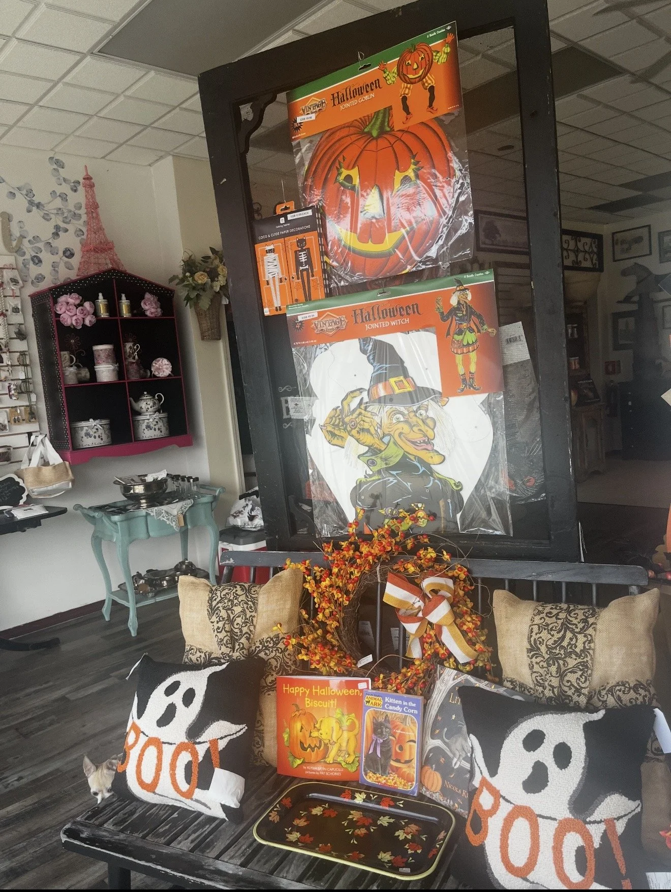 Fall-themed Halloween display with pumpkin and witch decorations on a black frame, surrounded by Halloween-themed pillows, books, and fall foliage in a store setting.