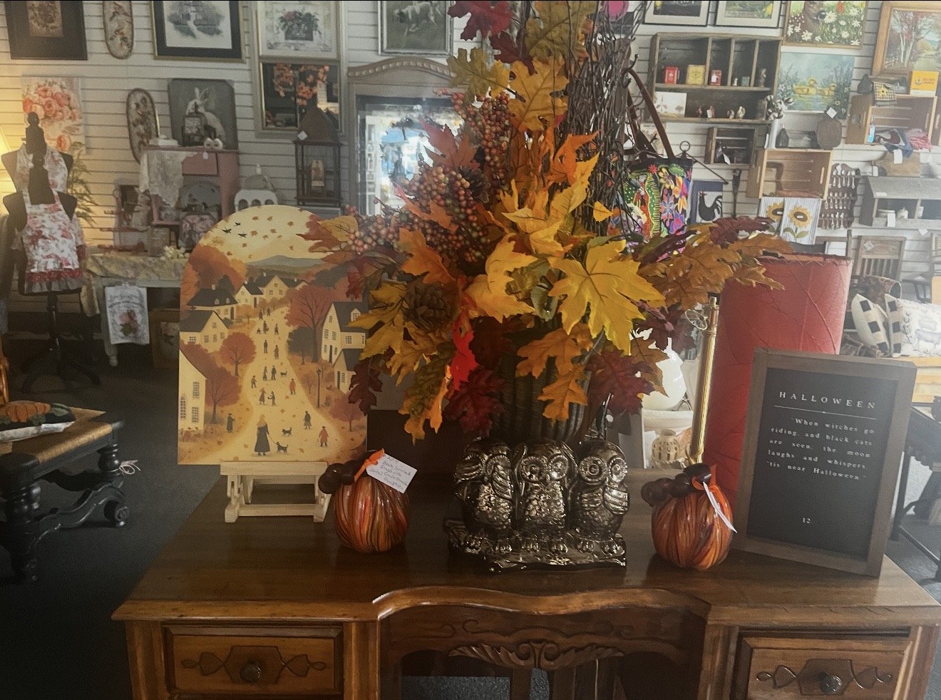 Autumn-themed display with a large bouquet of orange, red, and yellow leaves and berries in a black vase on a wooden table. There are two small pumpkin decorations on either side of the vase, and a Halloween sign to the right. In the background, a pi