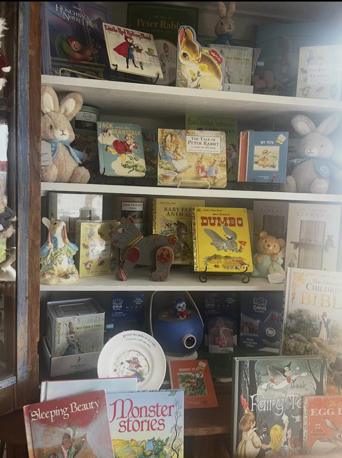 Bookshelf with children's books, stuffed animals, and toys, including titles about Peter Rabbit, Dumbo, and fairy tales, as well as plush rabbits and decorative items.