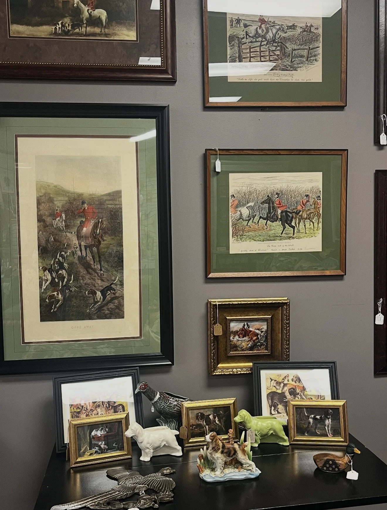 A display of framed hunting dog and hunting scene artwork on a wall, alongside small dog figurines and decorative plates on a black table.