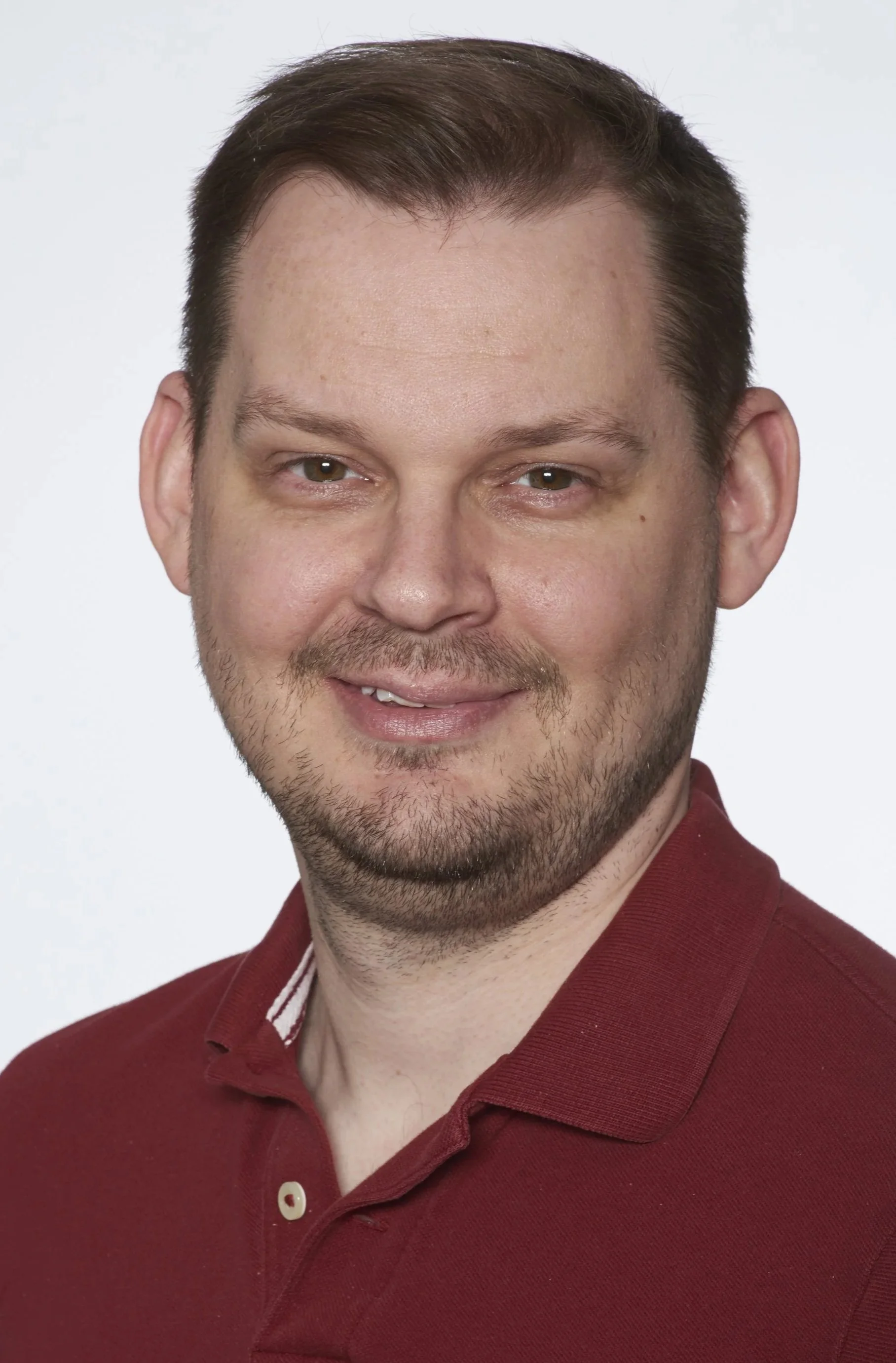 Close-up portrait of a smiling man with short brown hair, light facial hair, and wearing a maroon polo shirt with a white and maroon stripe on the collar, against a plain white background.