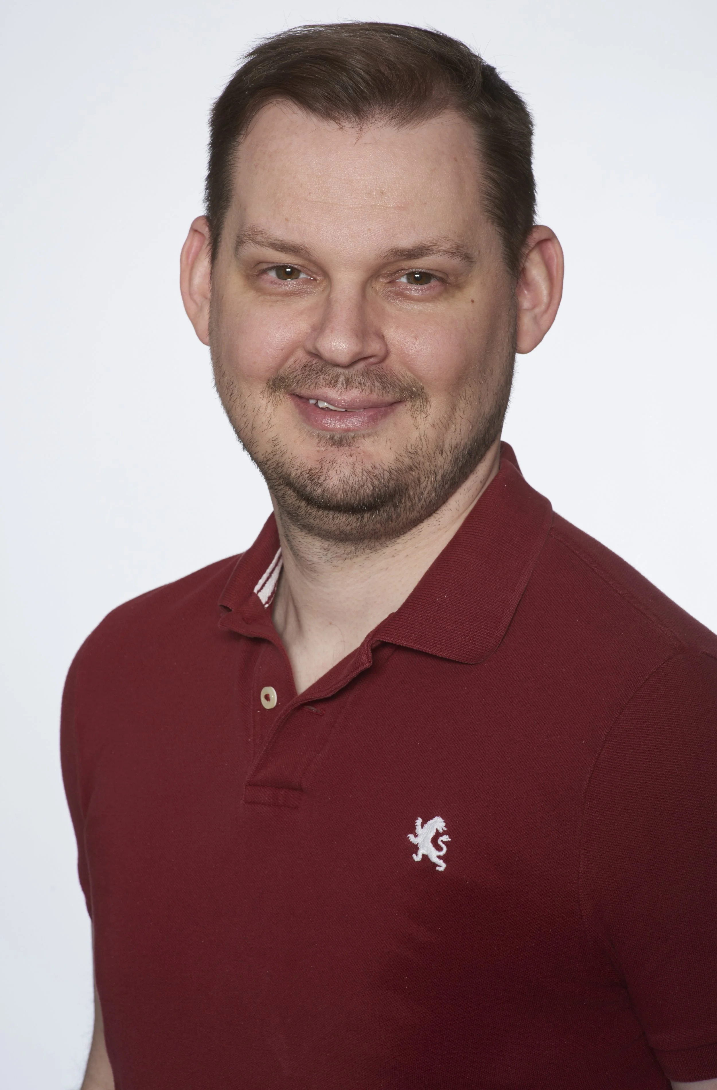 Headshot of a smiling man with short brown hair, a beard, wearing a maroon polo shirt with a lion logo on the chest, against a plain light background.