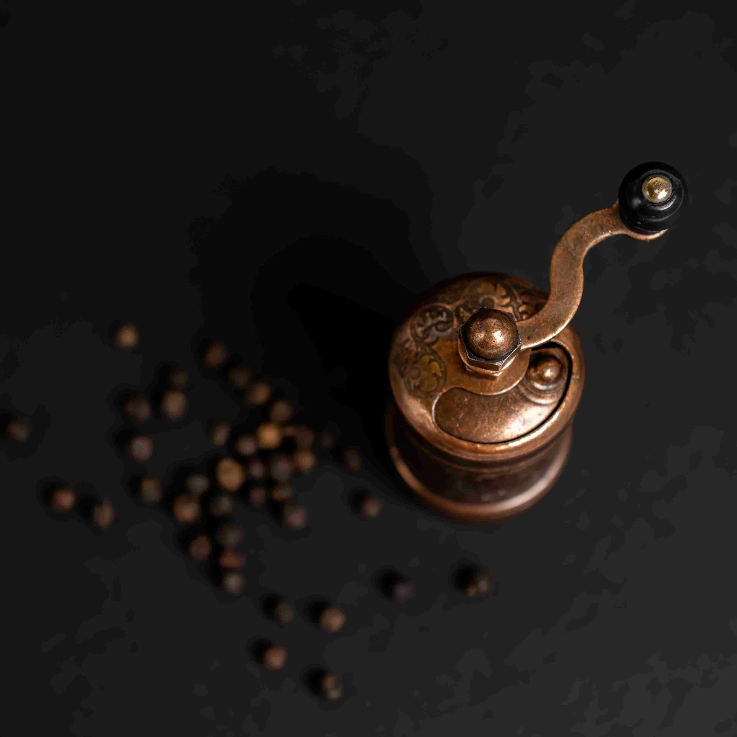 Antique copper pepper grinder on black background with peppercorns
